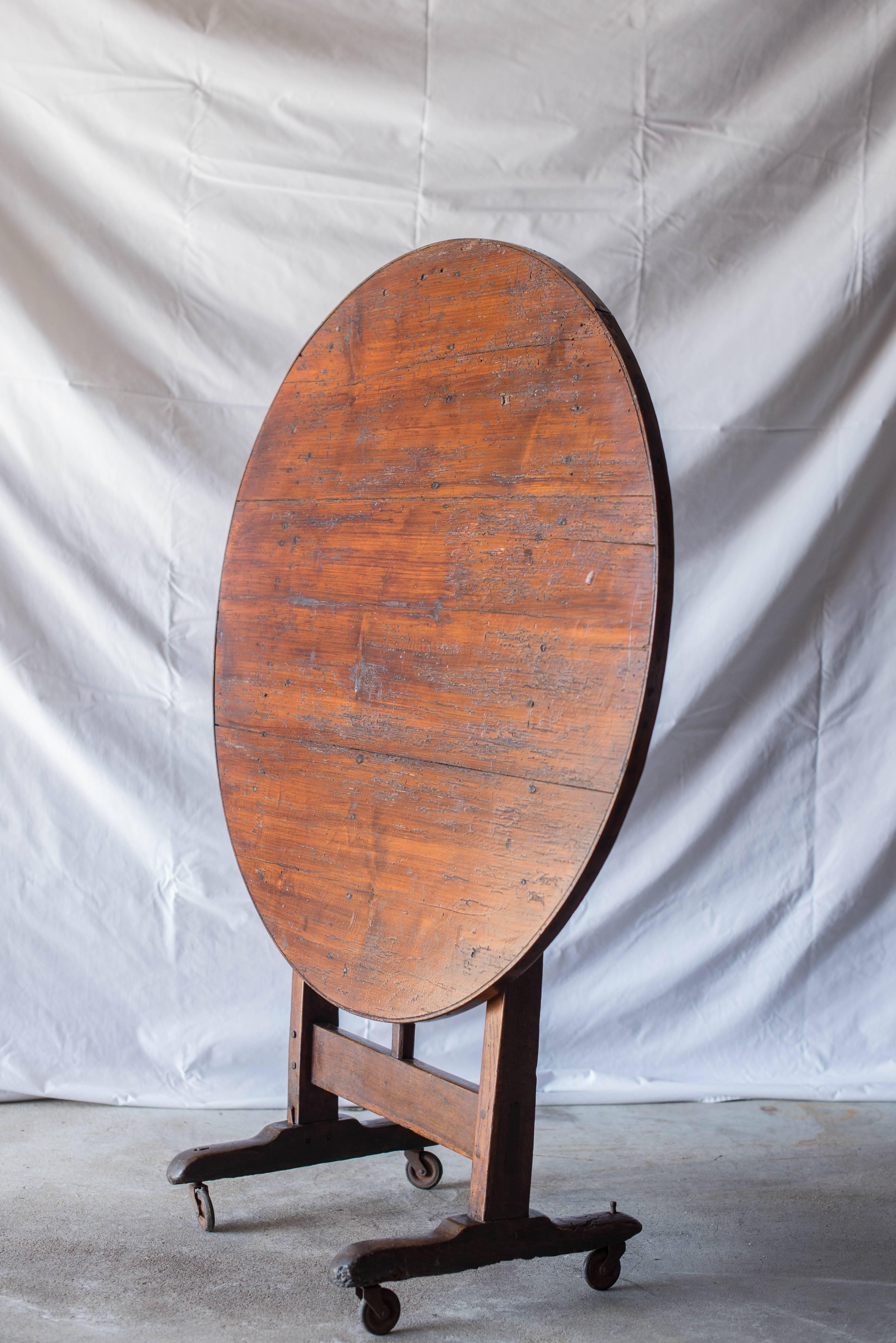 This tilt-top wine tasting table has an oak base and bentwood banding around the top. The top does not appear to be oak and can be positioned vertically for storage against a wall. The base has sledge feet and sits on casters.