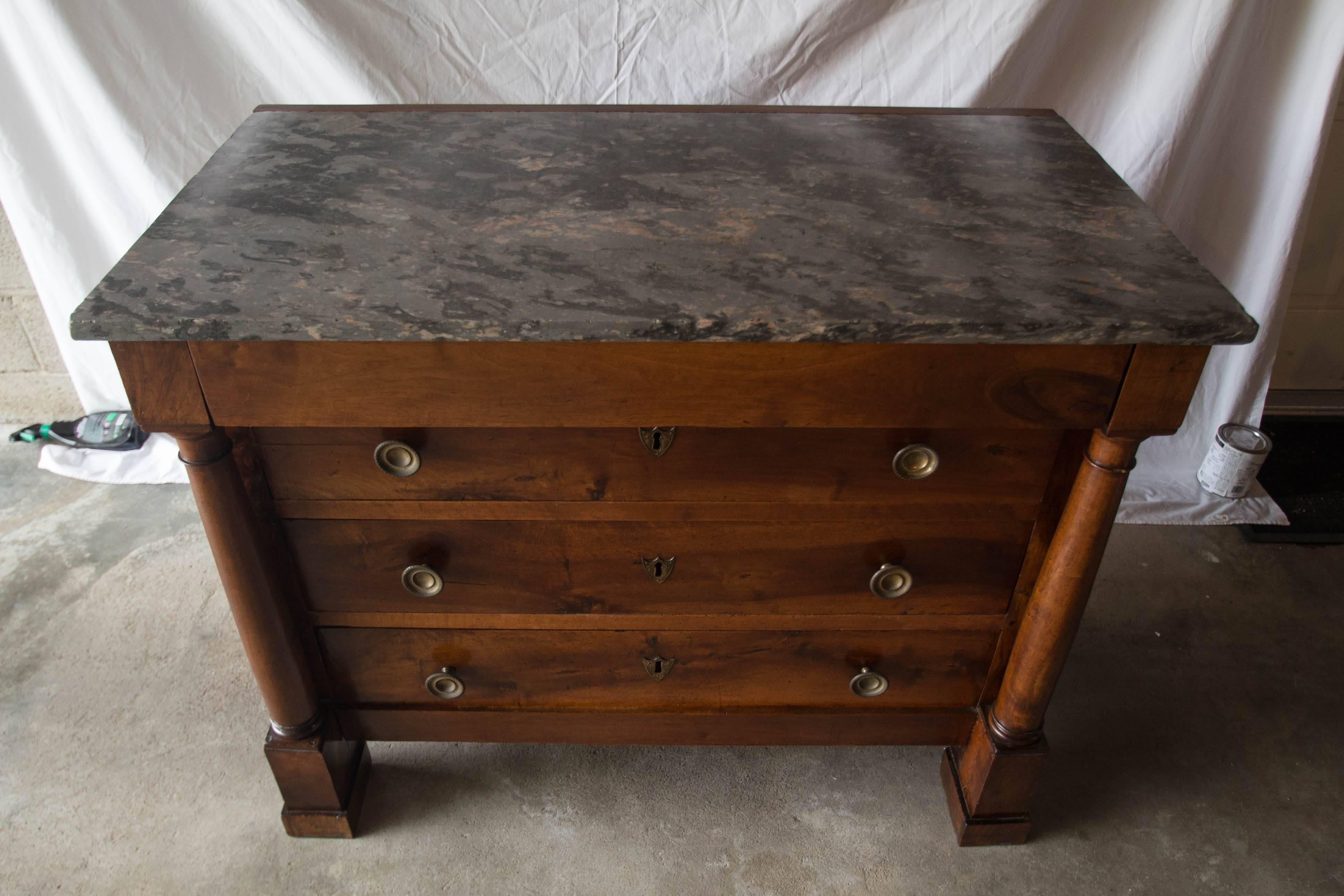 This lovely walnut commode in the Empire style features column pilasters with squared legs. The patina is rich and showcases the original brass hardware and key escutcheons. The marble top is different shades of gray with hints of terracotta.