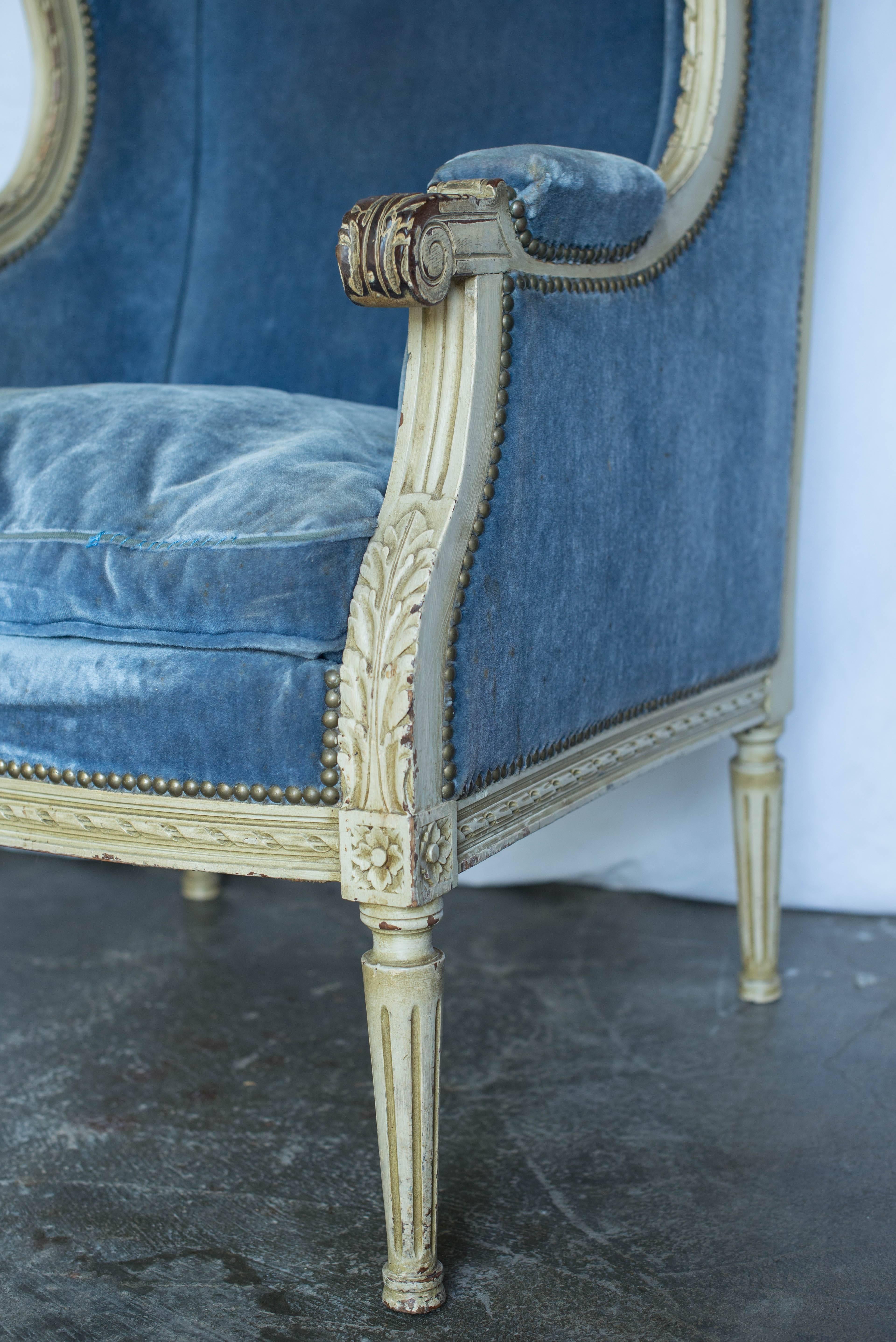 This pair of 19th century French Louis XVI style bergere chairs features beautiful carvings and a lovely painted cream finish. The painted finish on the arms has been worn revealing stained wood beneath. The blue upholstery is stained and needs to