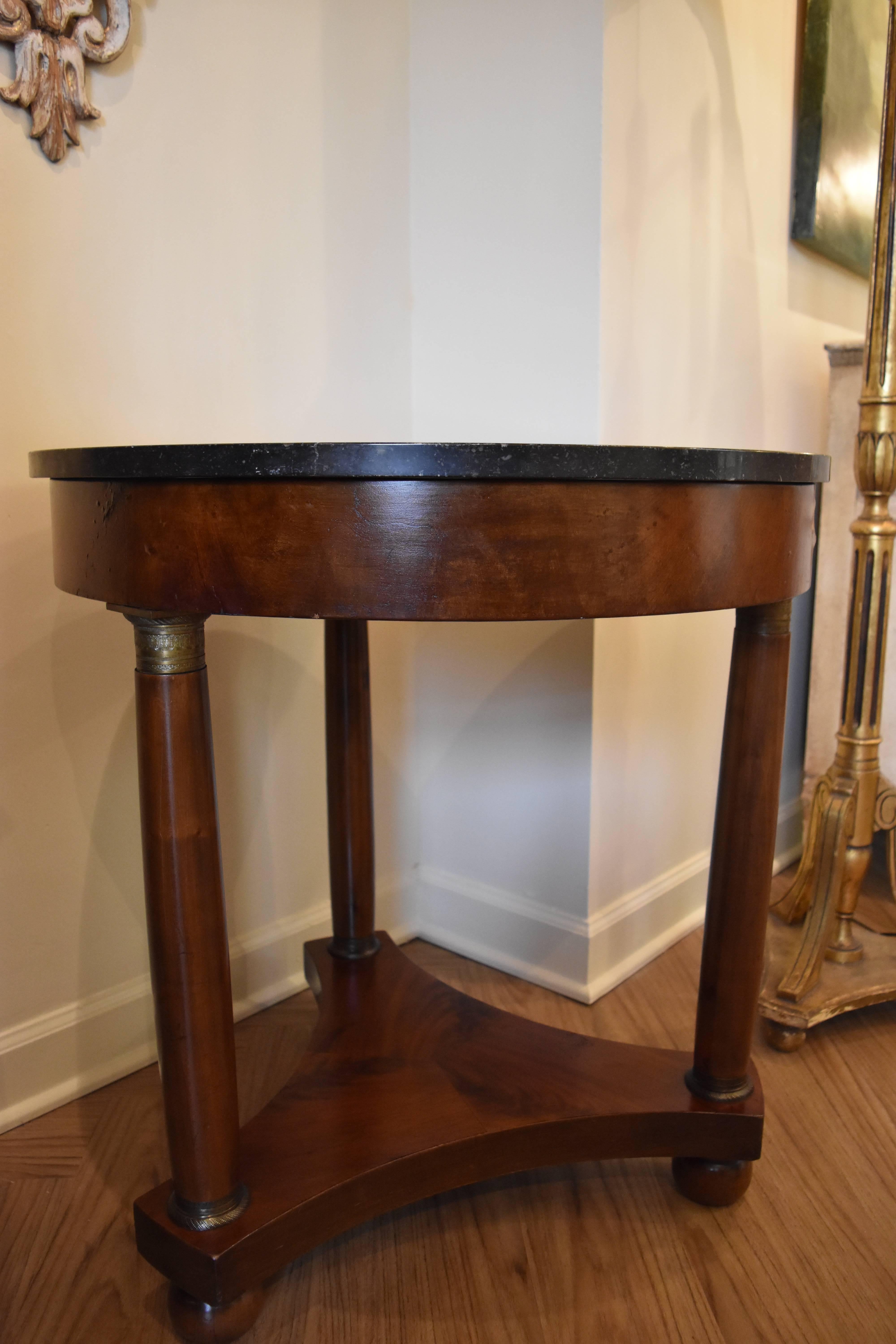 This walnut Gueridon table has a tripod base and bronze detailing at the top and bottom of each leg. The marble is black with several minor scratches. The veneer around the apron has several bubbles as shown in one of the photos.