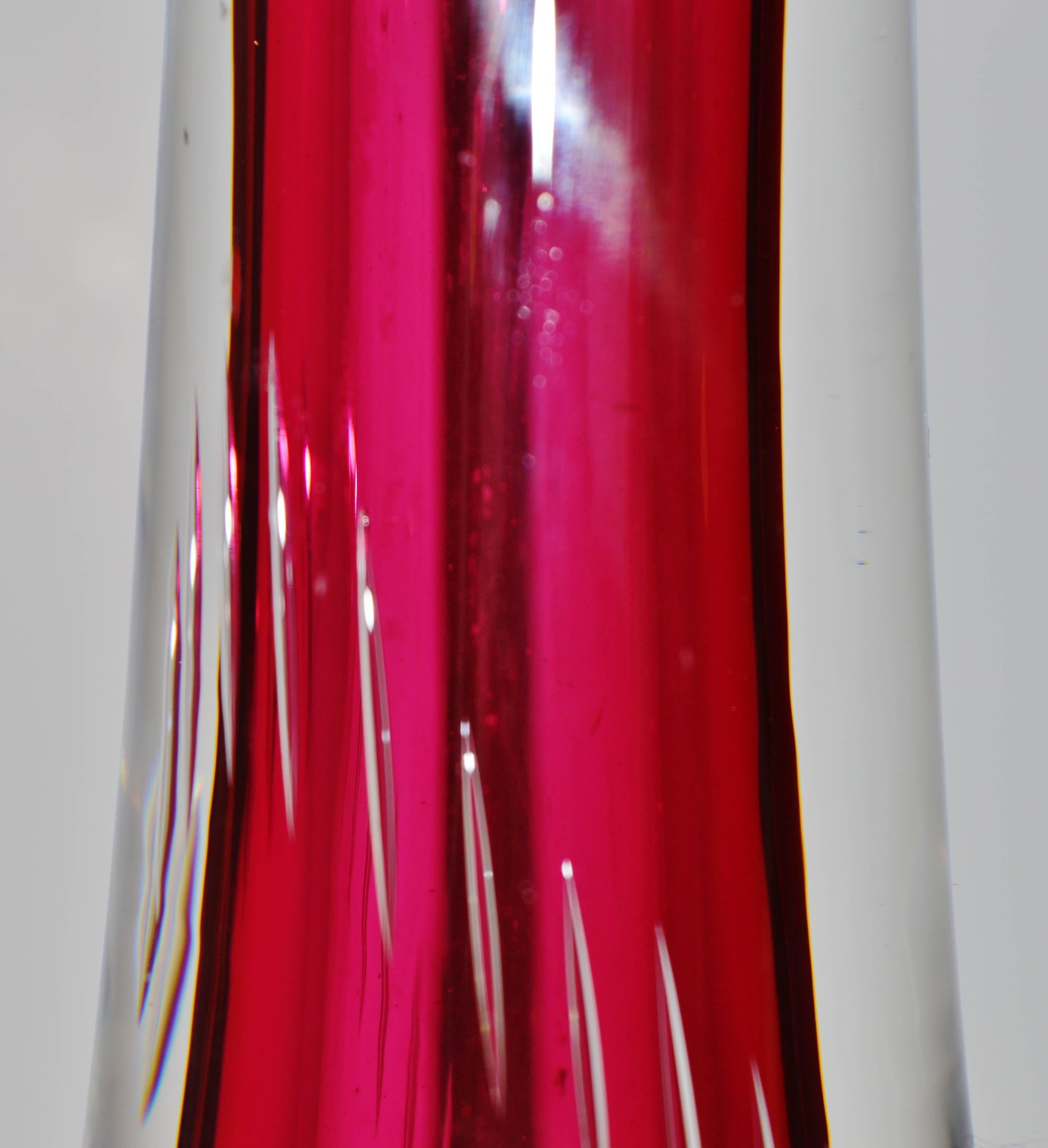 This lovely Kosta Boda vase in cranberry and clear features the controlled bubble, but the vase was elongated after the bubbles were inserted, such that the bubbles in the middle are elongated. Angled mouth and in excellent condition. Signed Kosta