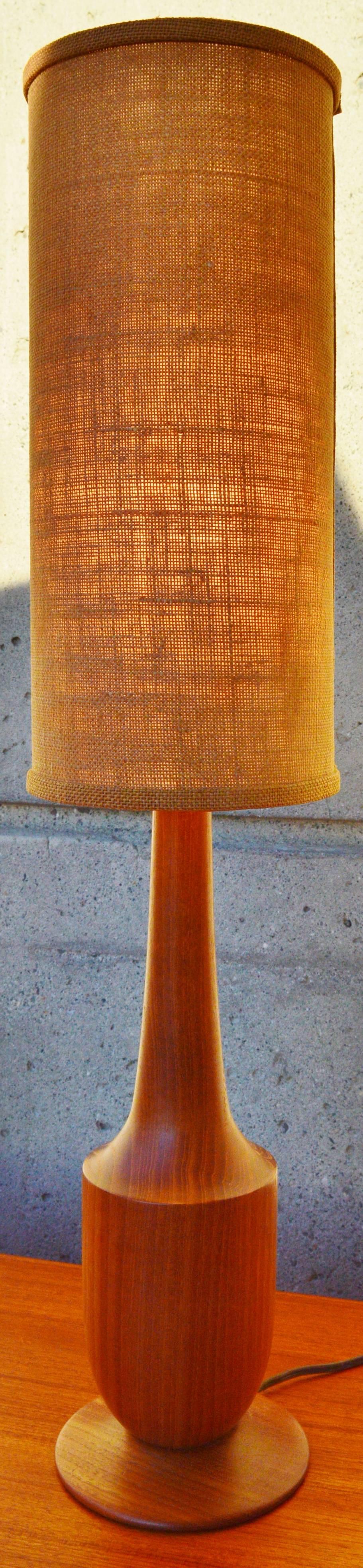 This Danish modern solid teak table lamp has gorgeous grain patterns throughout the sculptural form and disk base. Complemented beautifully by the original tall and thin jute cylindrical lamp shade. In excellent condition and with a nice rich