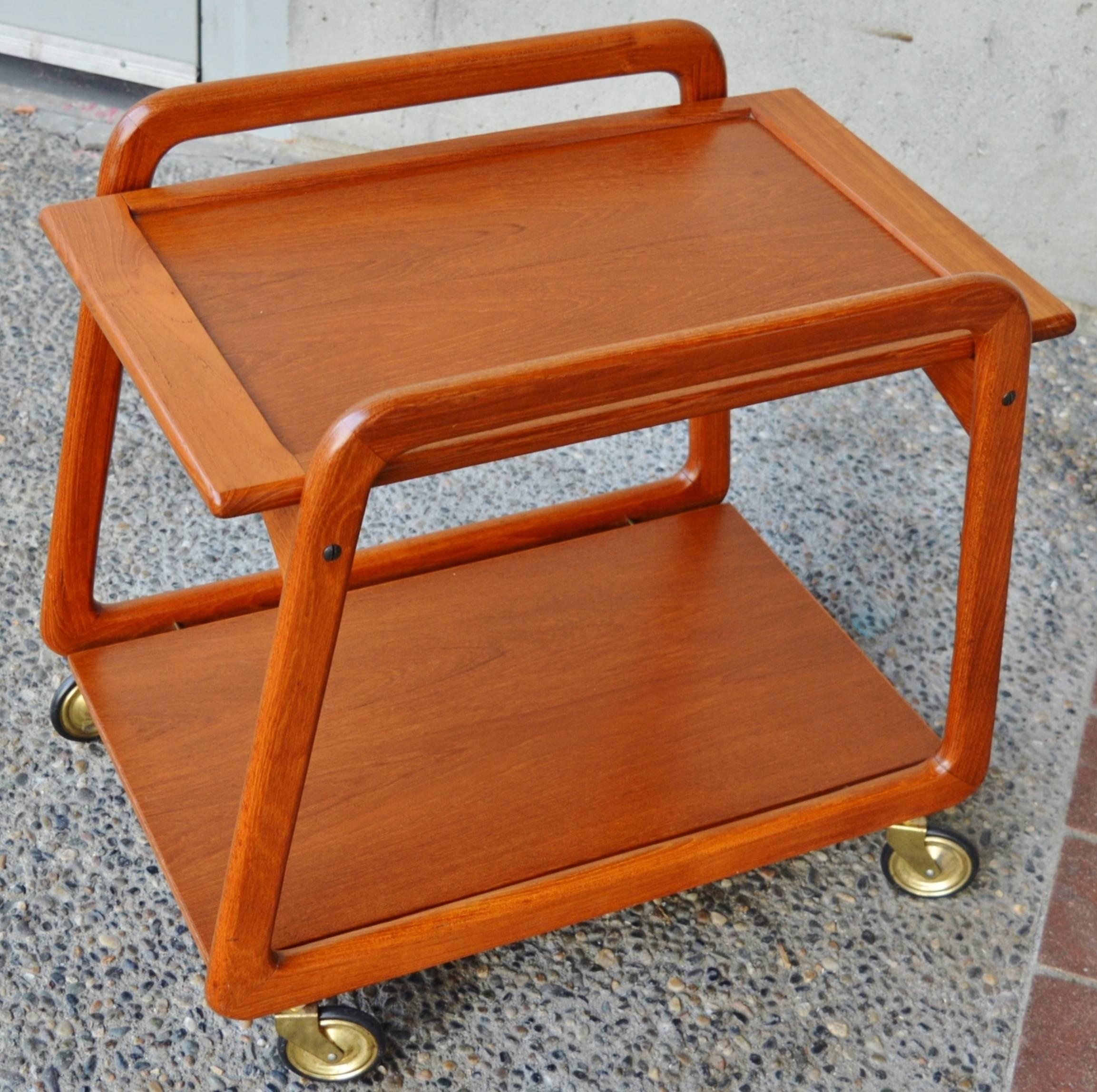 This super fun Danish modern teak bar cart or tea trolley has awesome lines! It features trapezoidal shaped sides, arced upper braces that support the two side’s tray top, a lower floating shelf held by brass pins, and Classic brass skinny 50s