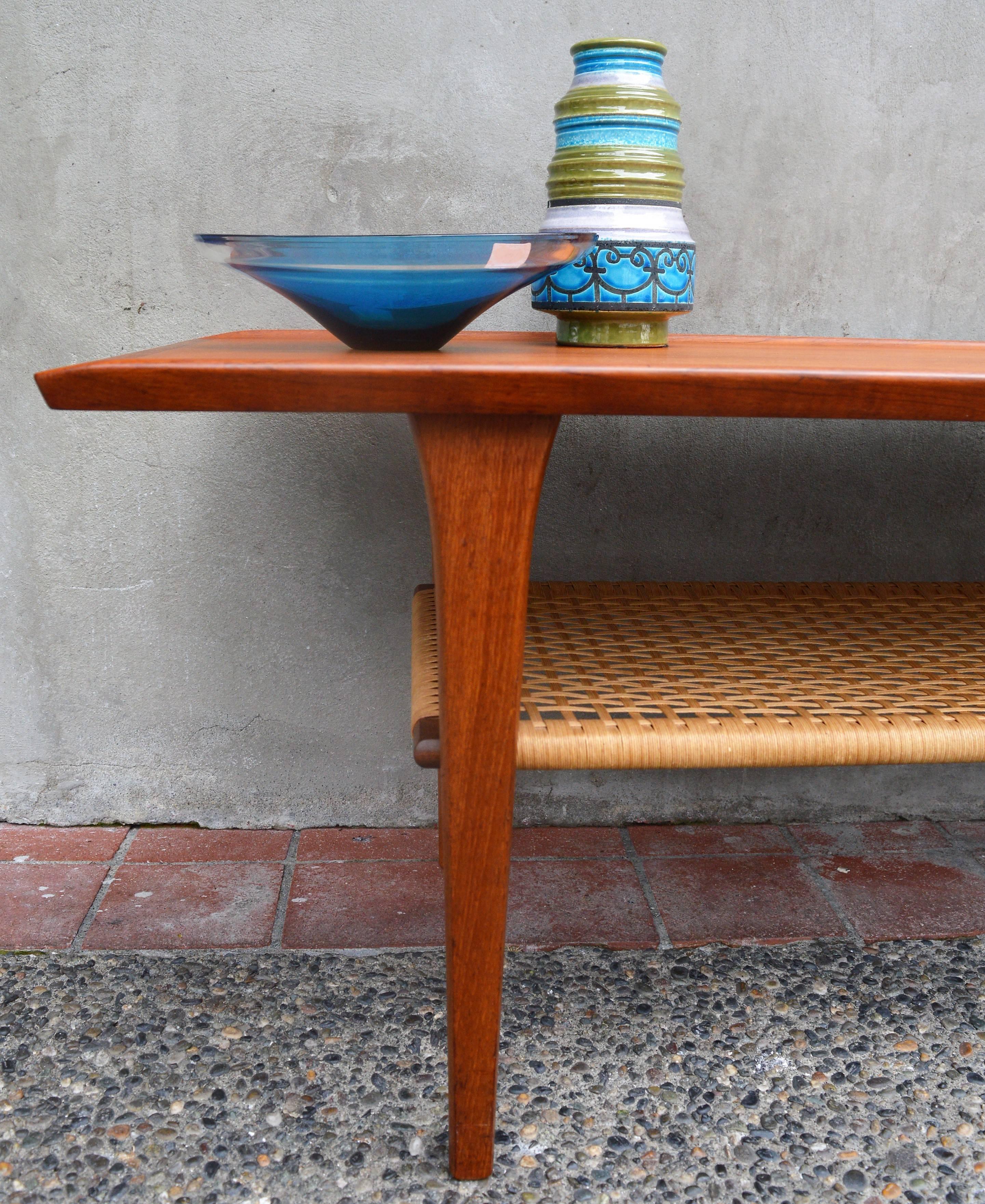 This stellar, more compact Danish modern teak coffee table manufactured by Mobelintarsia in the 1960s. Note how the legs flare below the table top which has flared top edges. The lower shelf is woven with a unique multi strand caning that wraps the