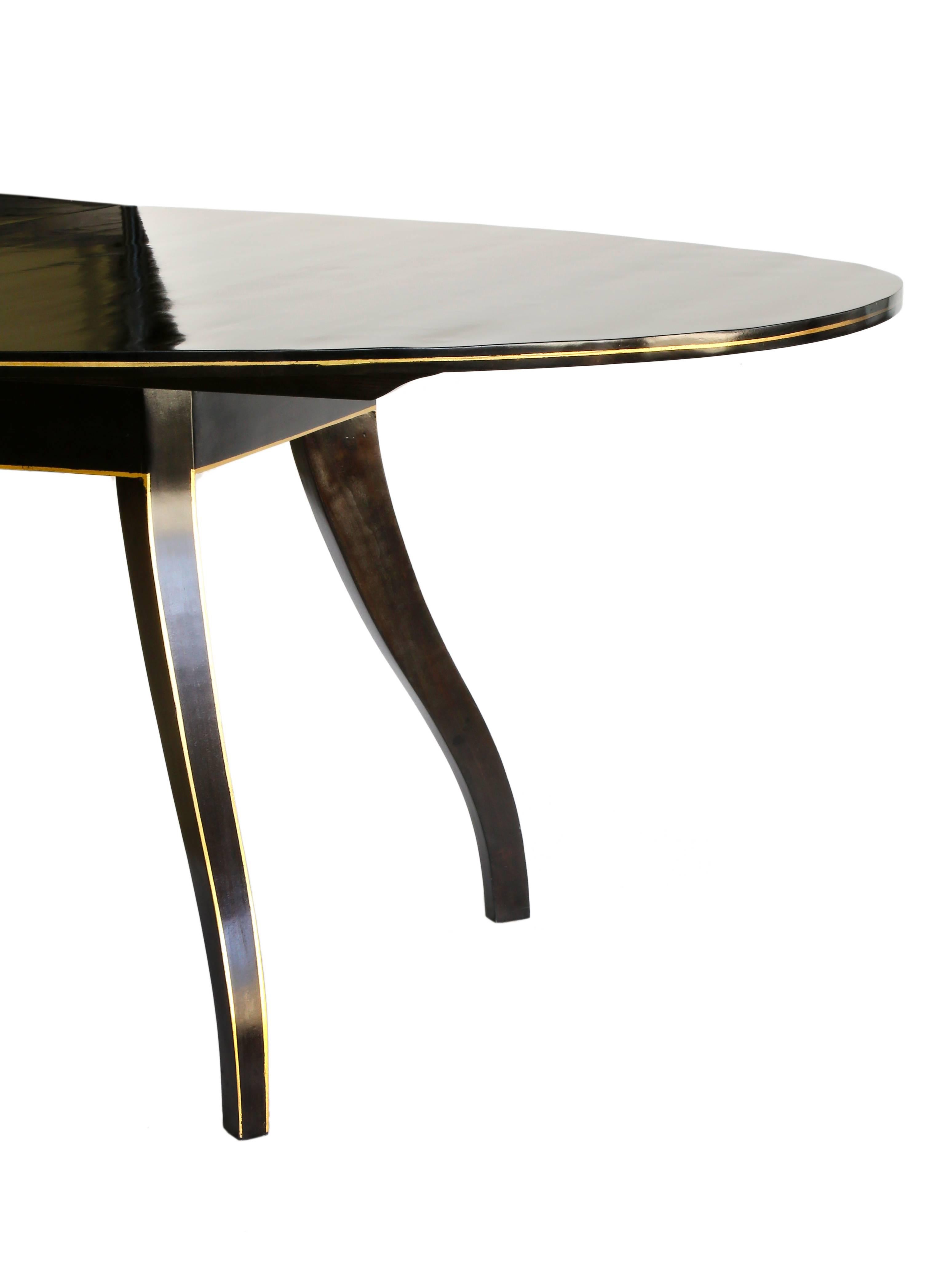 An extension dining table designed by Phoebe Howard in dark ebony stained wood. The table features Regency style spider legs with a painted 23-karat gilt banding detail on the edge of the leg and along the edge of the table. This table style is no