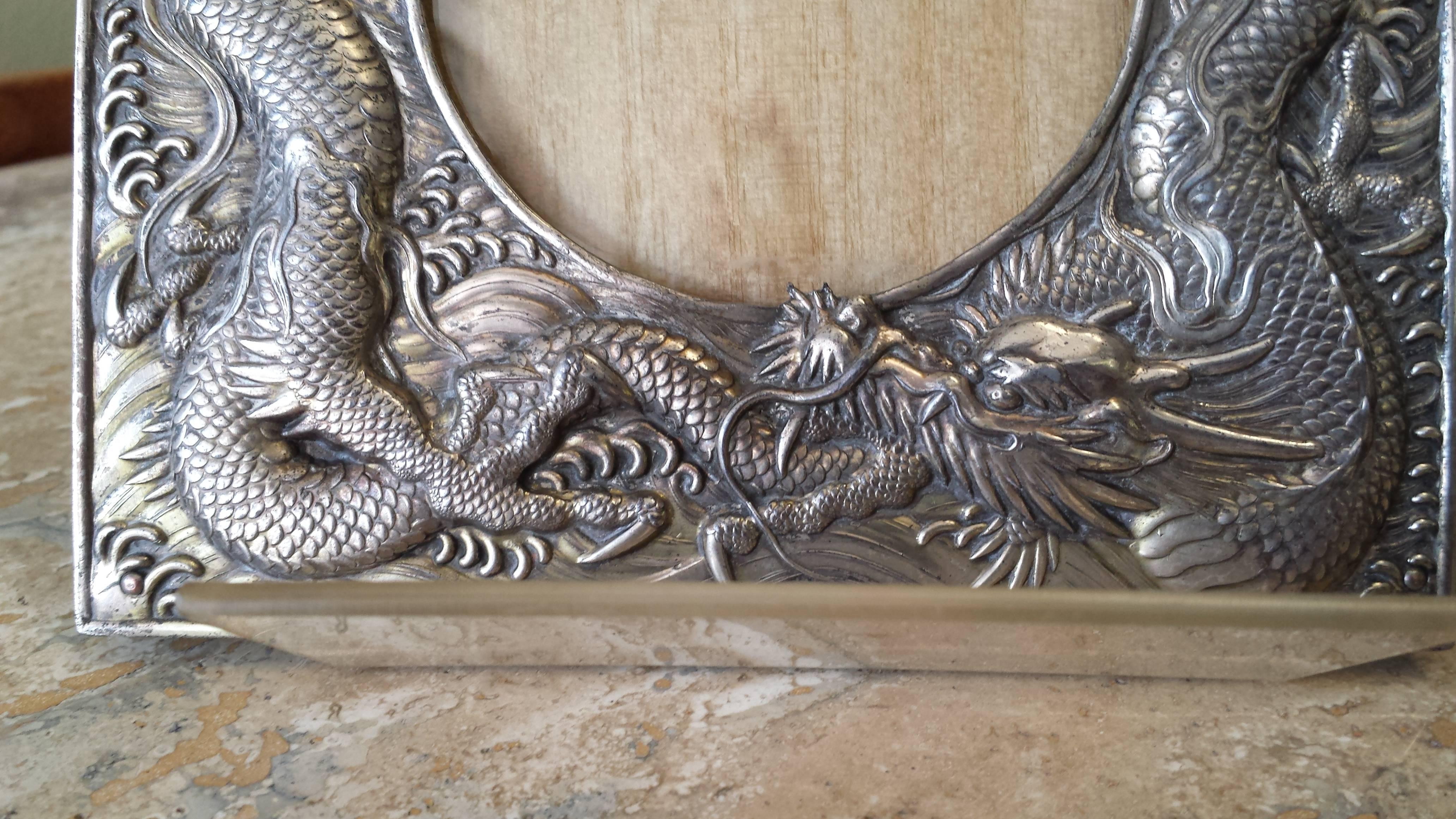 Stunning raised double dragon picture frame in a silver/gilt old finish, circa 1900-1910. The frame is flanked by two dragons and flanks a round picture hole, the back is in original vinyl covering and wire. The frame measures 5 7/8