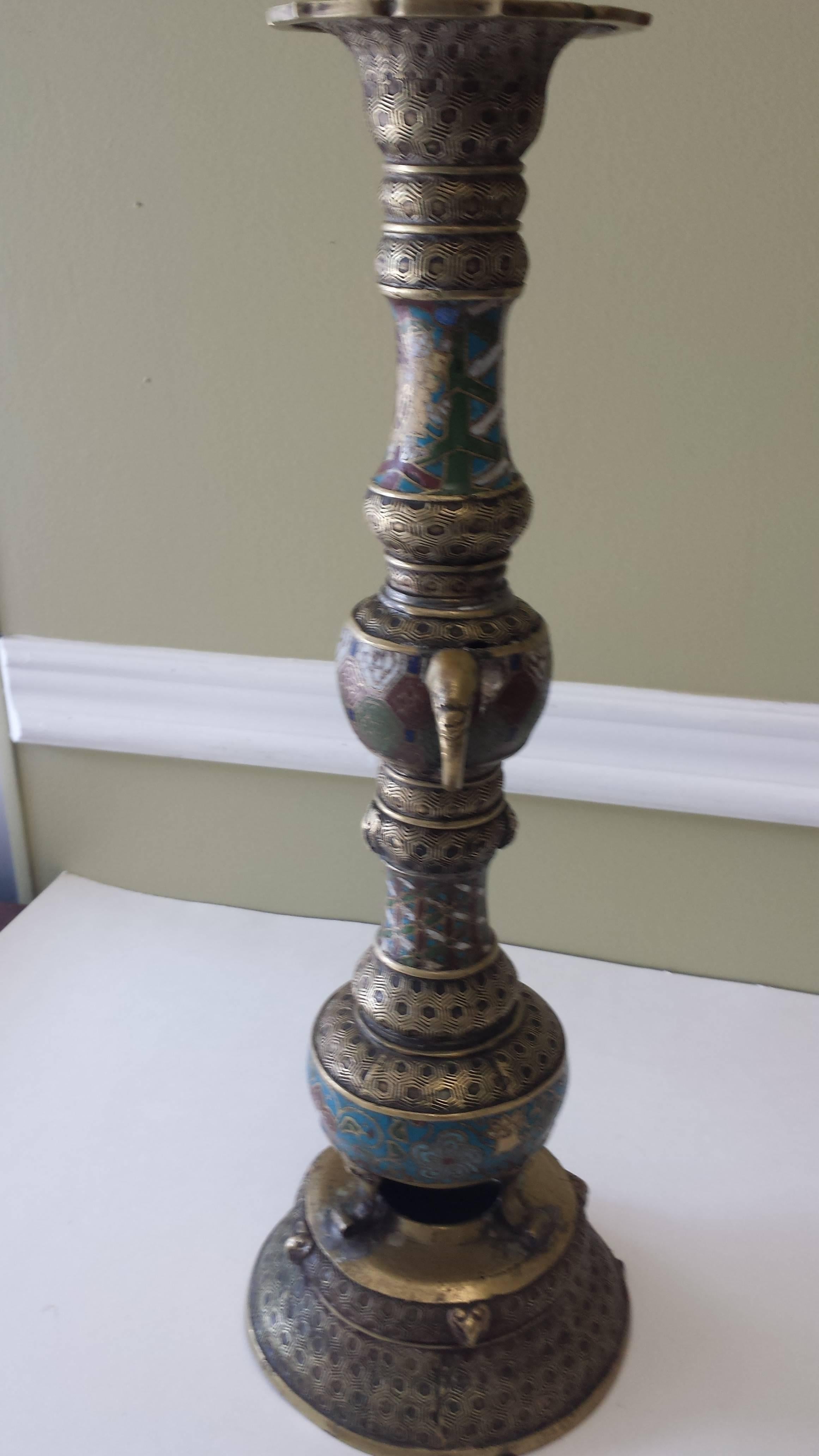 A bronze Champlevé Buddhist temple Altar candleholder, Meiji period, with stylized elephant head handles and Ram's heads decoration. The enamel work has some loss and there is a stress crack/worn split on the top section of the base, which can be