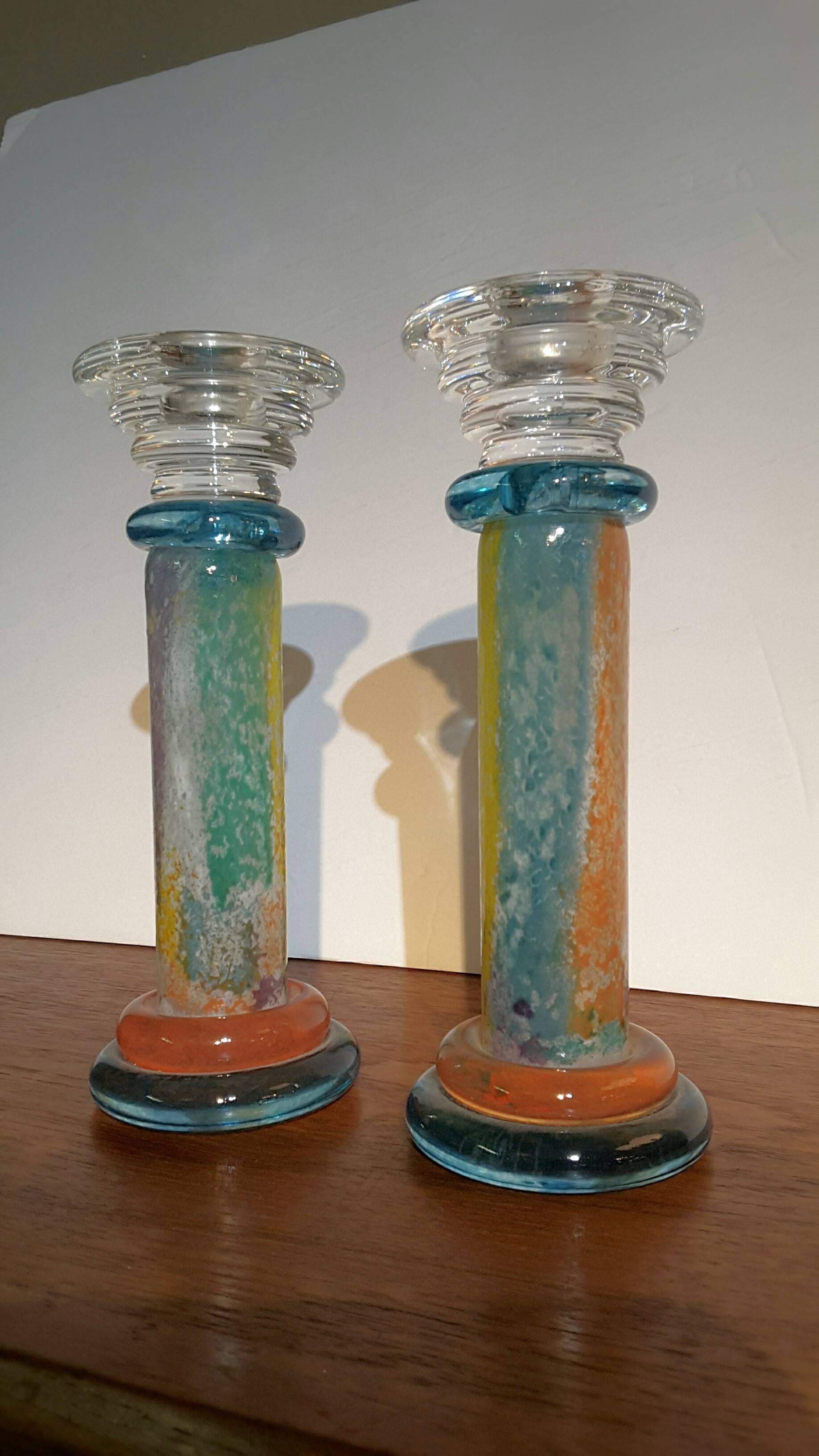 A pair of Kosta Boda art glass candlestick holders designed by Kjell Engman. The candlestick holders features a rounded blue base with a second orange/ red base on which stands a cylindrical column decorated in textured hues of yellow, blue, and