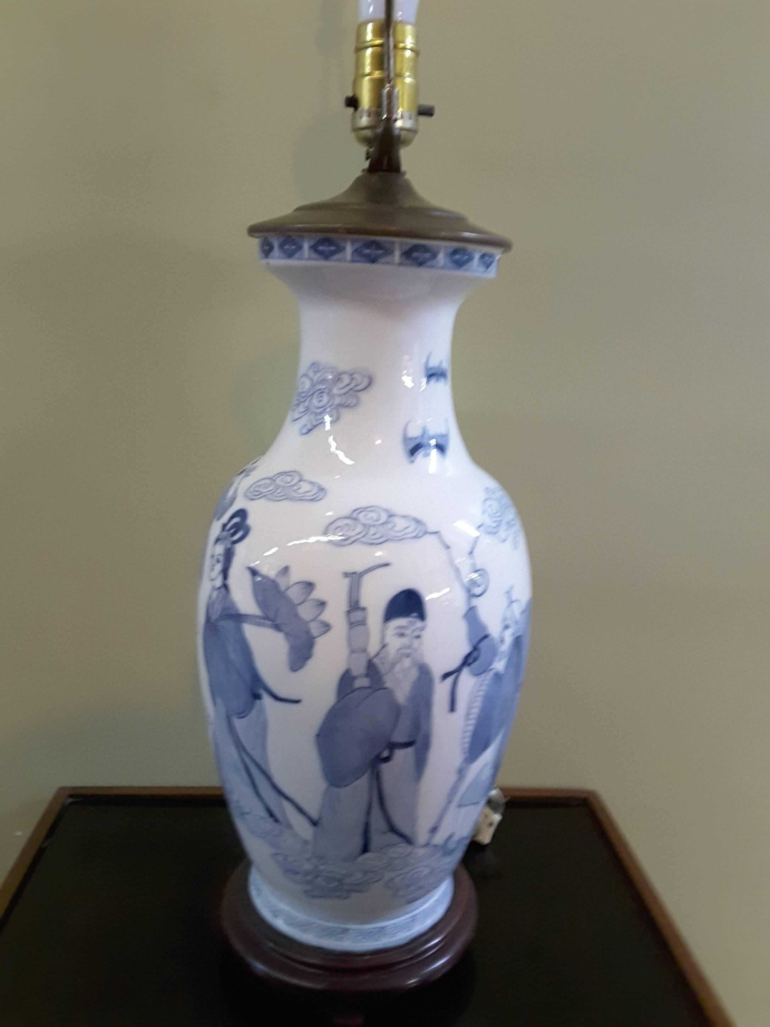 An Asian pictorial blue and white table lamp, the lamp is done in a typical blue on white background, there are eight different figures around the baluster shaped base, there are bats, clouds, figures and banded on the top and bottom. Mounted on