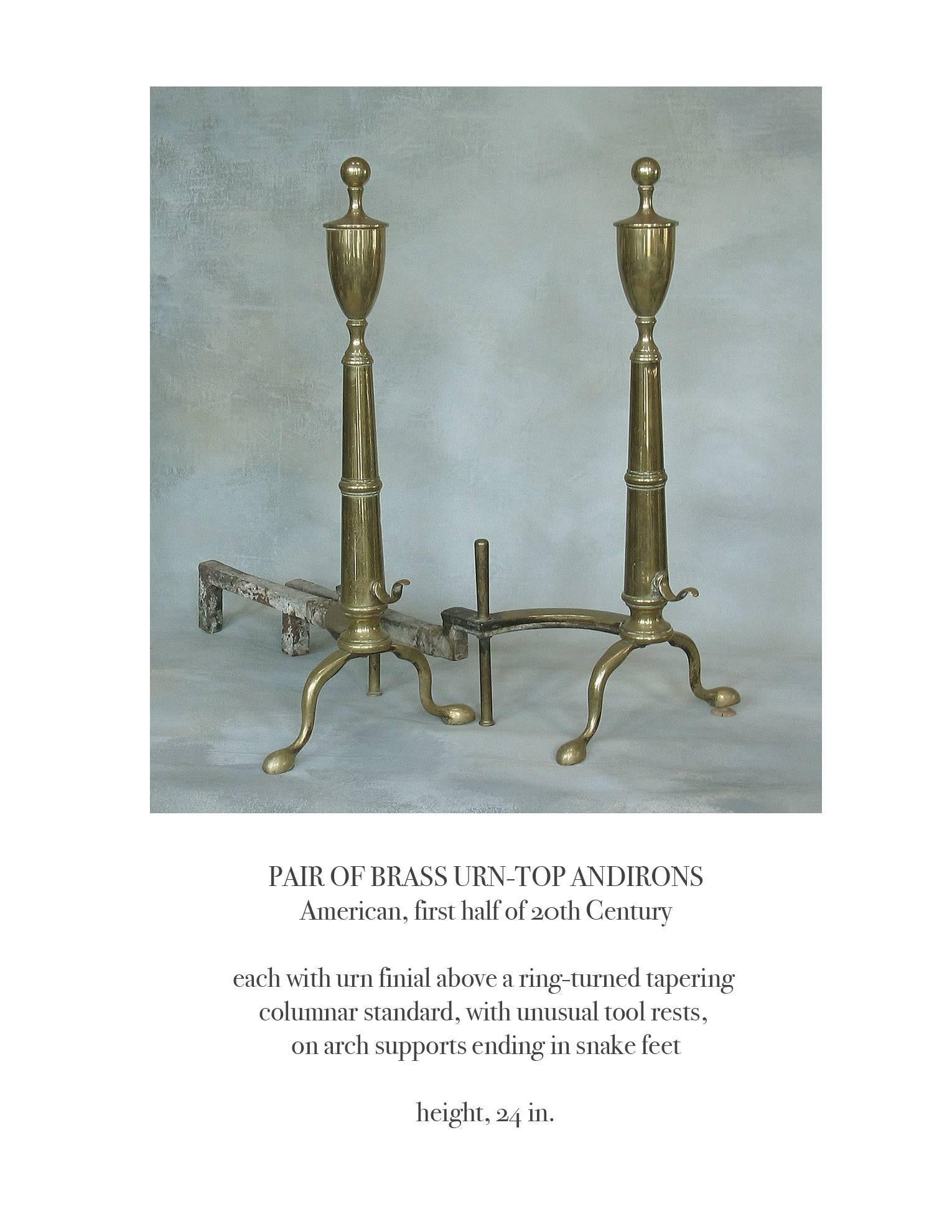 Pair of brass urn top andirons, American, first half of the 20th century, each with urn finial above a ring turned tapering columnar standard with unusual tool rests on the front, on arched supports ending in a snake pad foot. The andirons measure
