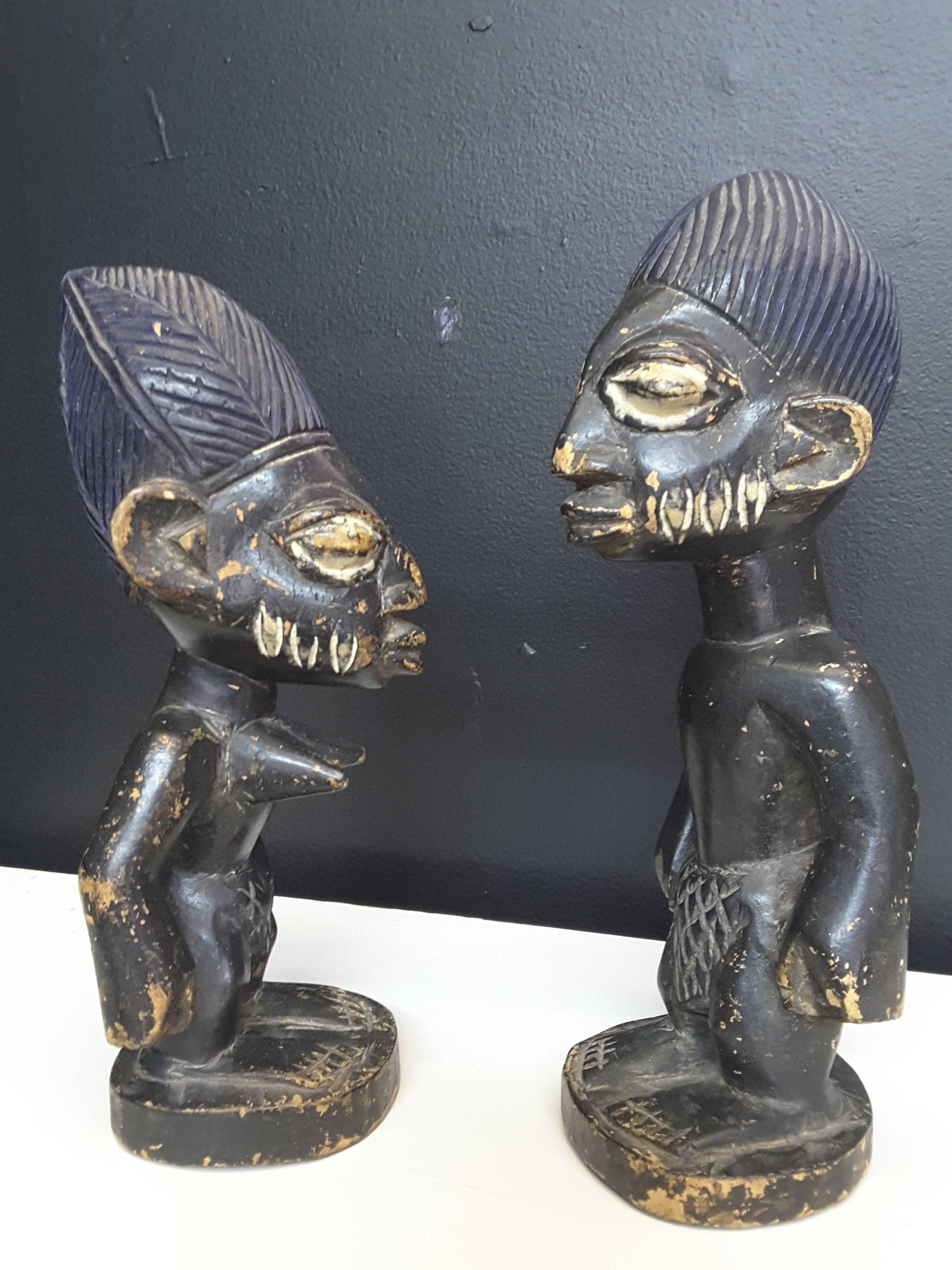 A pair of Yoruba Culture Ibeji (Nigeria) statues, a male and female carved figural statues with exaggerated features. Both figures have elongated blue headresses, frontal loin clothes, done in a dark black finish with white highlighted eyes and