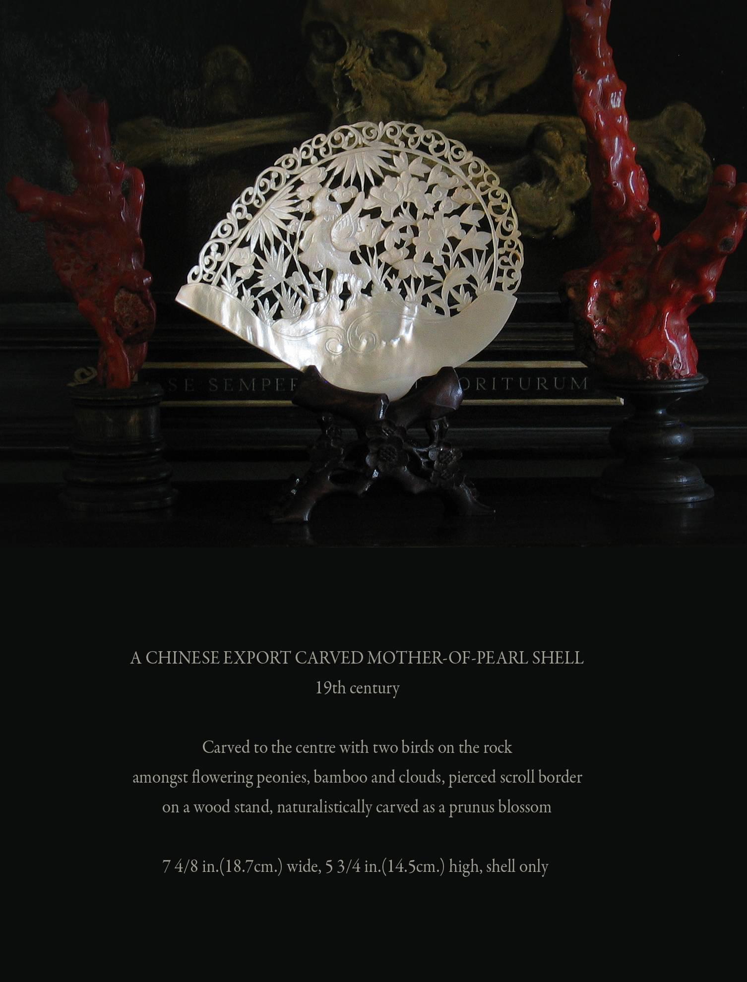 A Chinese export carved mother-of-pearl shell, 19th century, carved to the center with two birds on a rock amongst flowering peonies, bamboo and clouds, pierced scroll border on a wood stand, naturalistically carved as a prunus blossom.
The carving