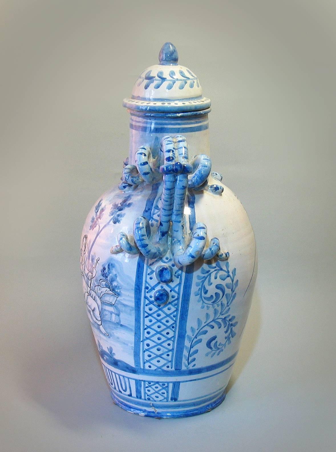 A large Italian blue and white Majolica covered urn/apothecary jar, Savona (Levantino Family), early 19th century, of baluster form with domed lid and twin snake handles on either side, decorated with a pastoral scene on either side and a large