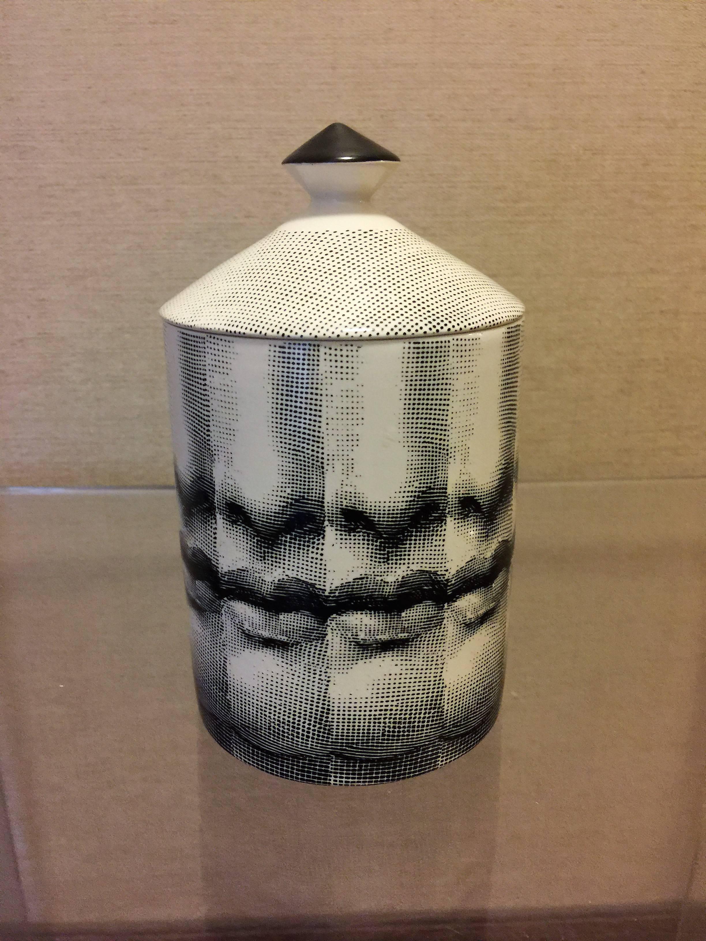 A more Modern ceramic cylindrical box that holds a scented candle made by Fornasetti decorated with repeating black and white sexy facial features images.