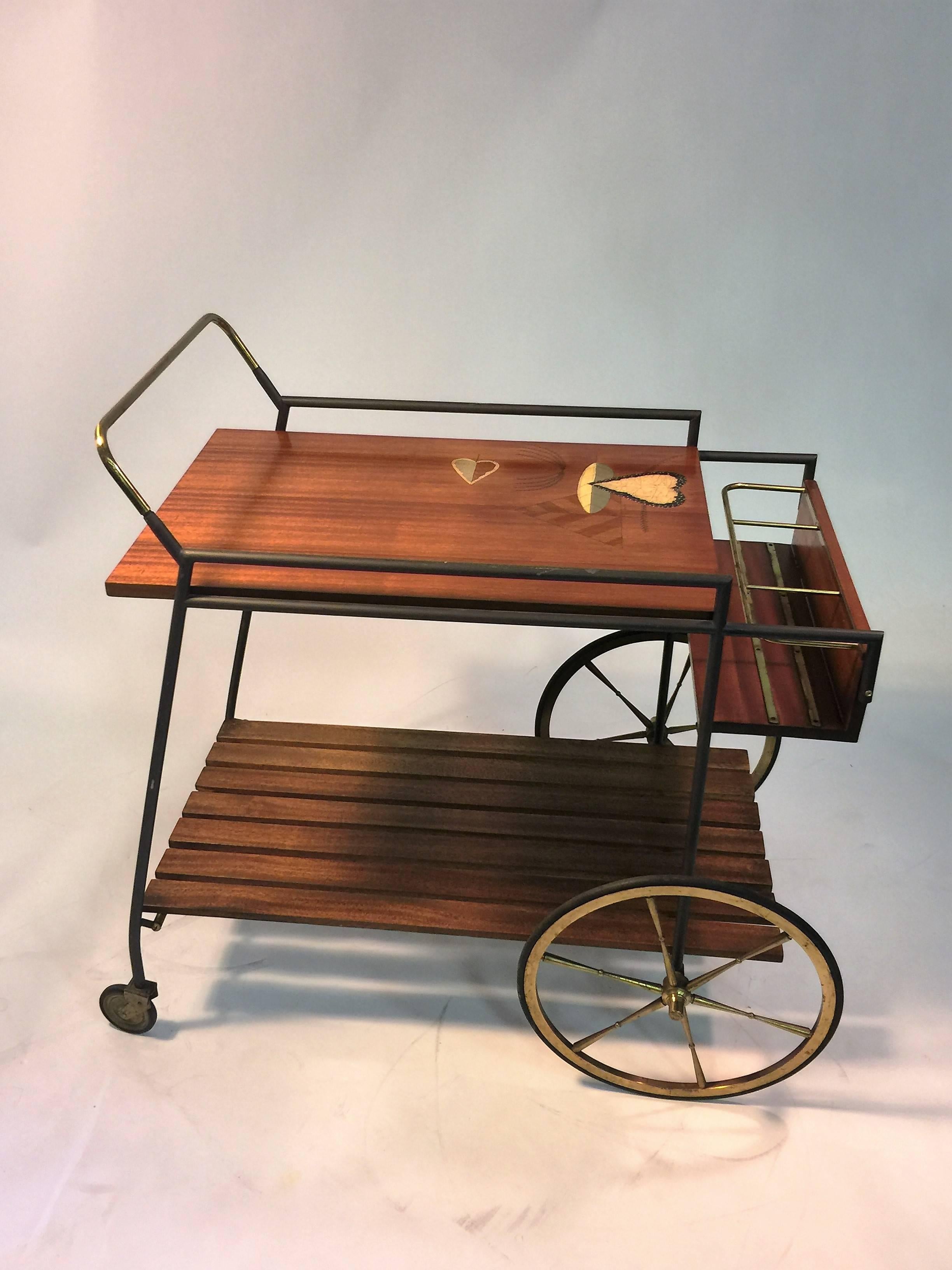 Interesting bar cart with beautiful modernist leaf design painted inlay with artists signature though Illegible to me. Slatted wood lower shelf and brass and wood bottle holder. Great large brass wheels and blackened iron frame with polished brass