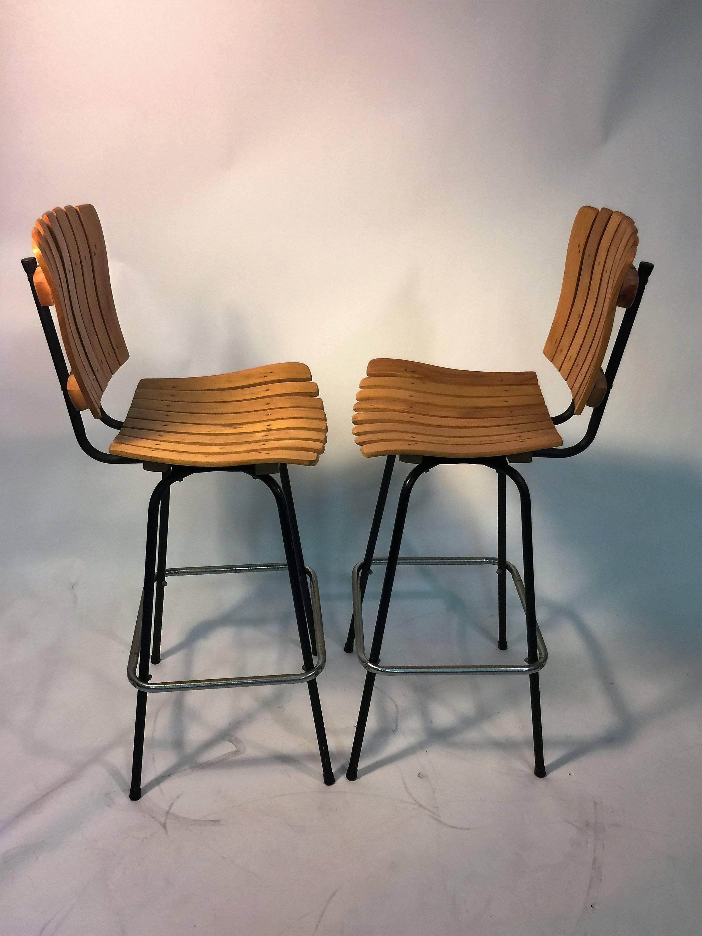 Set of four slatted wood design bar stools with black iron swiveling bases and chrome tubular surround foot rest. The black iron base measures at 16
