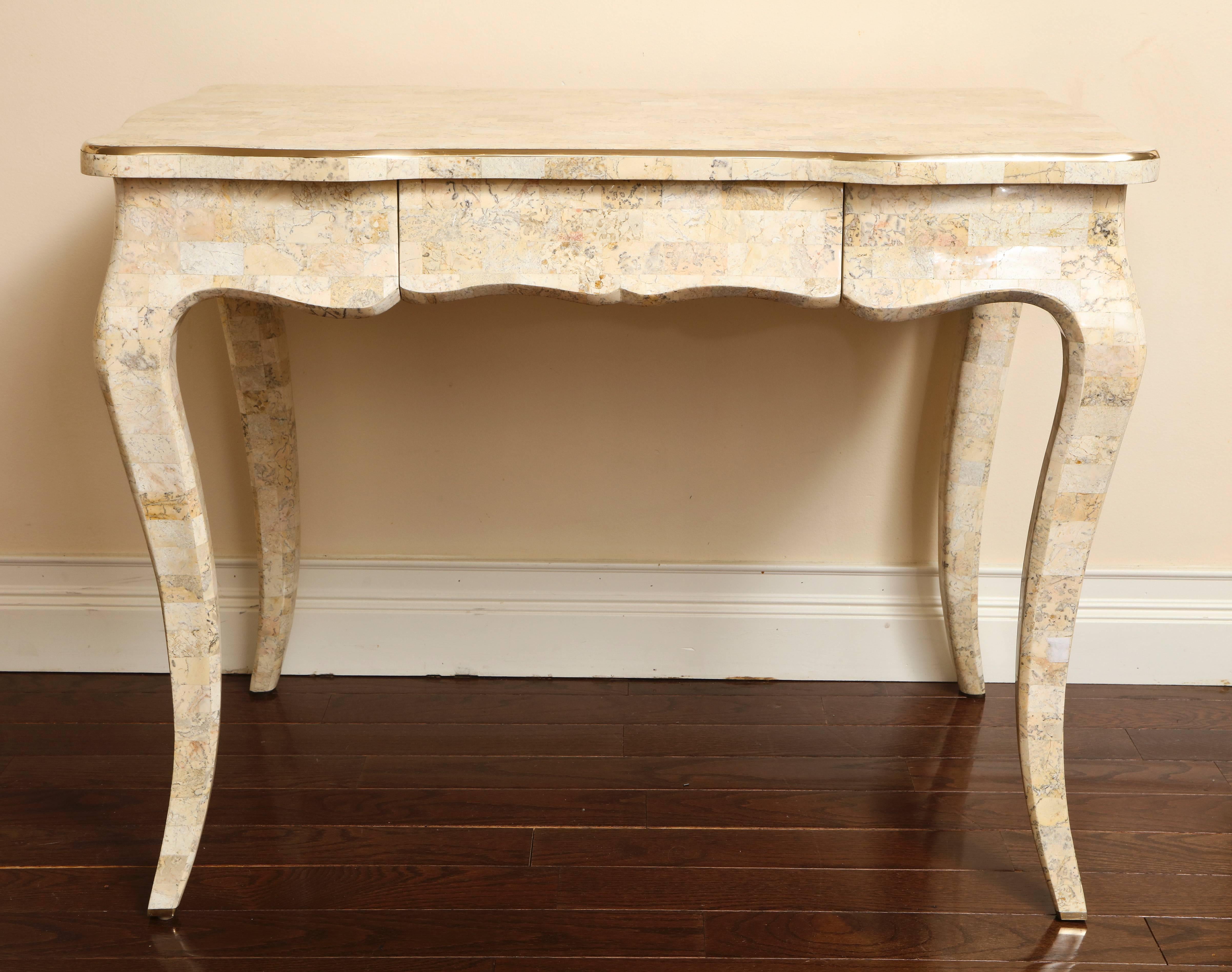 A Maitland Smith tesselated stone vintage desk with brass trim on the outer portion of the top. Of Rococo style; with scalloped apron and elegant cabriole legs terminating in brass sabots. There is a single drawer in the front with a signed brass