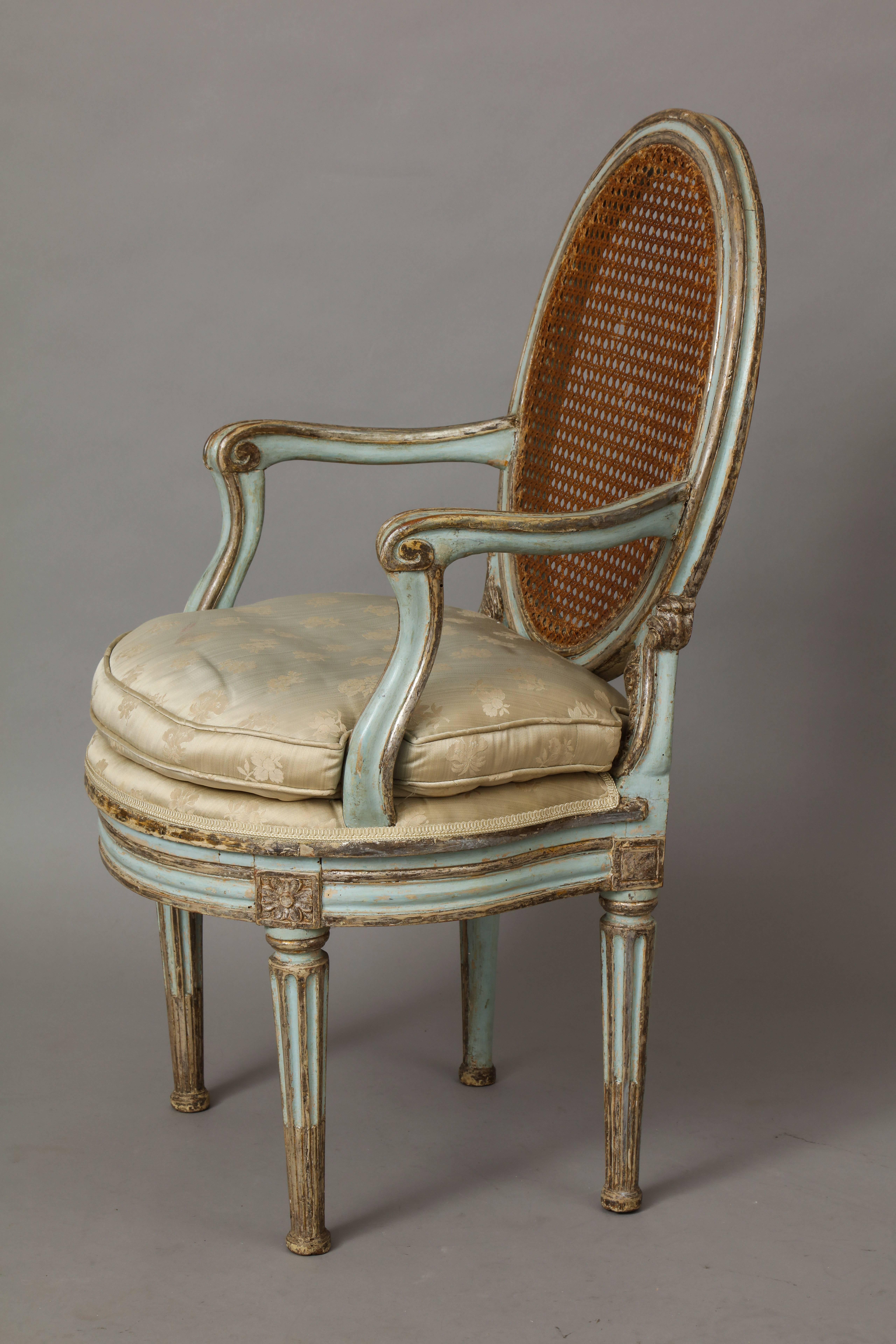 An original Louis XVI period Venetian painted and Mecca silver gilt armchair; with oval caned back, joined by scrolled armrests above an upholstered seat cushion above a molded circular apron with carved rosettes; tapered and fluted painted and