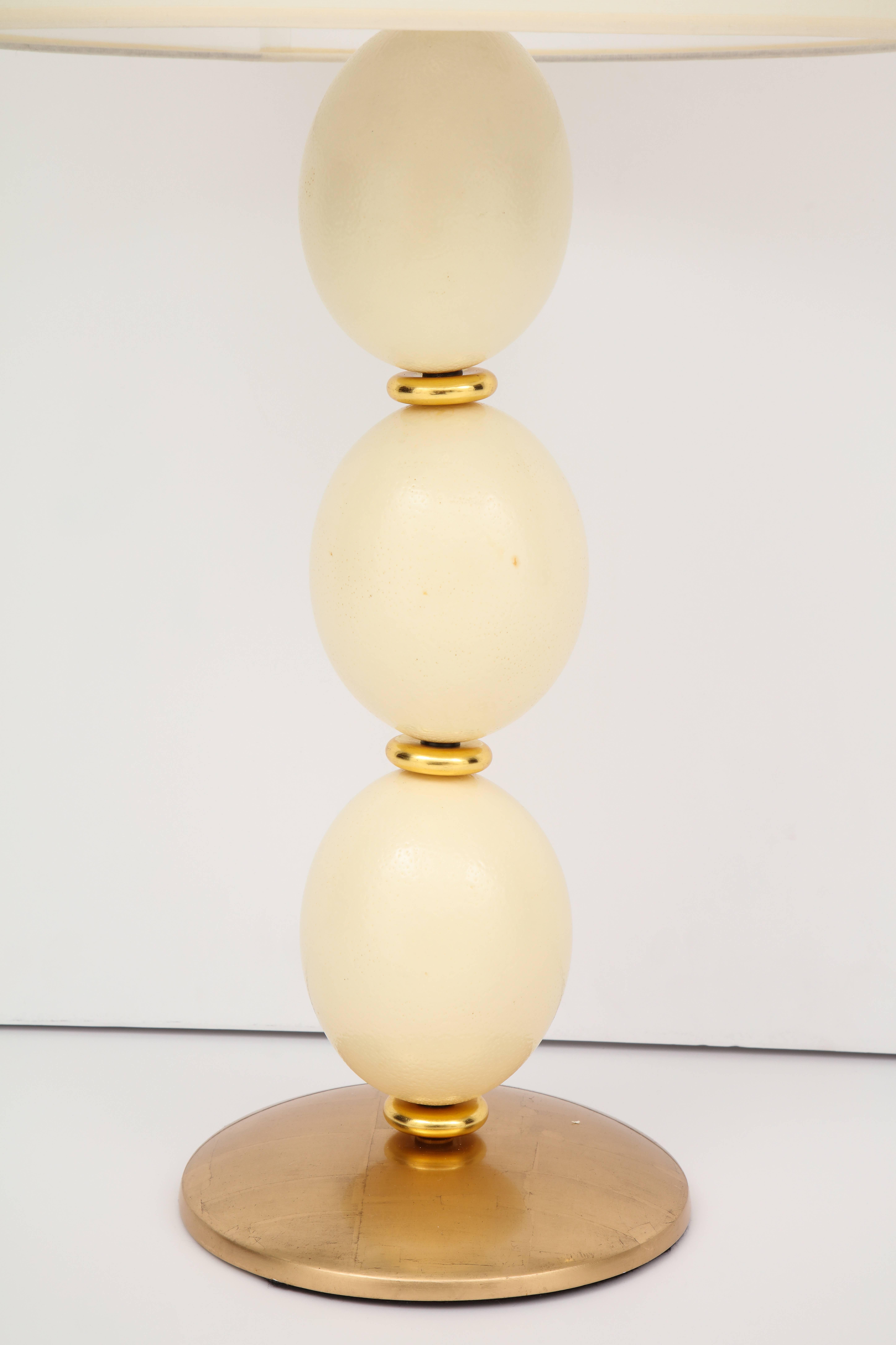 Une lampe de table faite sur mesure en bois doré sculpté et en œuf d'autruche. La base est dorée à l'or rose mat 28 carats, les anneaux en or jaune bruni, le fleuron en laiton doré et en os sculpté.
Abat-jour inclus si désiré.
Taille : 33