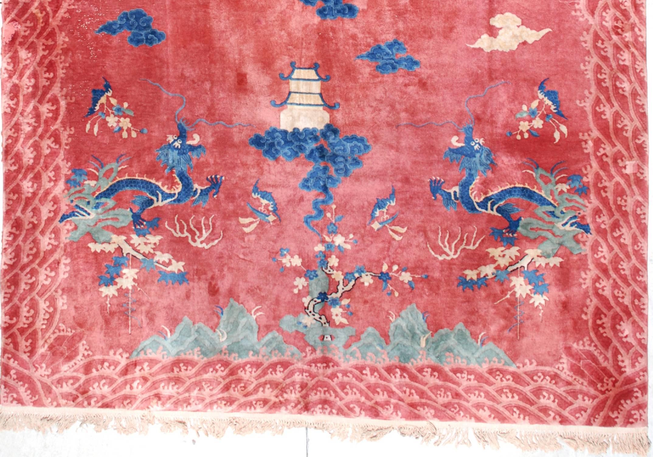 This Art Deco Chinese rug (inv 6761) from circa 1930, measures 8‘10” x 11’9”. It has a strawberry red to dusty rose ground with a five toed dragon in ivory and two shades of blue coming out of a rockery and it has a tree motif in each corner. There