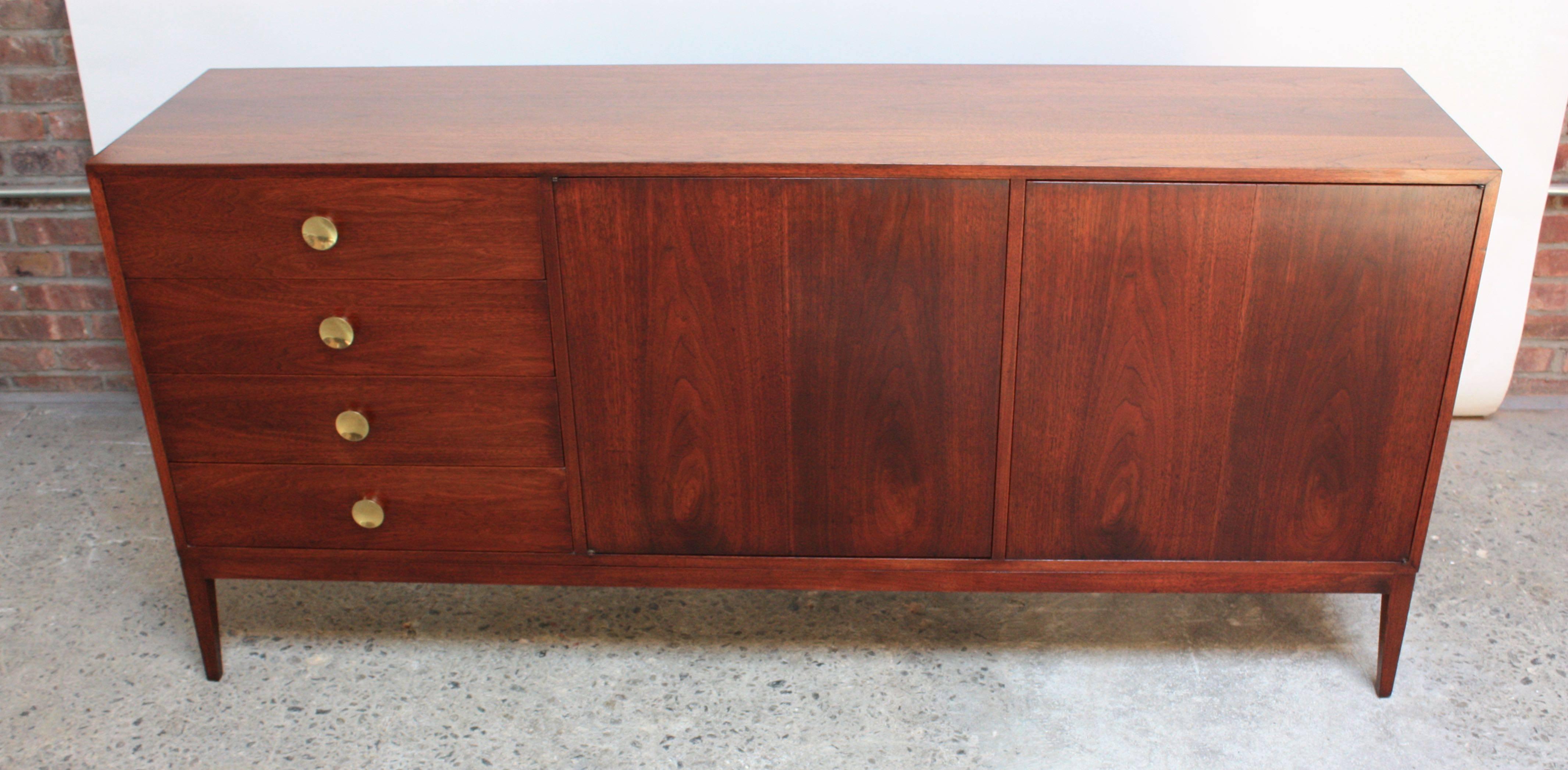 This exquisite credenza in walnut features four drawers with circular brass pulls and two bi-fold doors that open to reveal an additional eight drawers. This versatile piece offers sufficient storage for use as a dresser in a bedroom or a credenza.