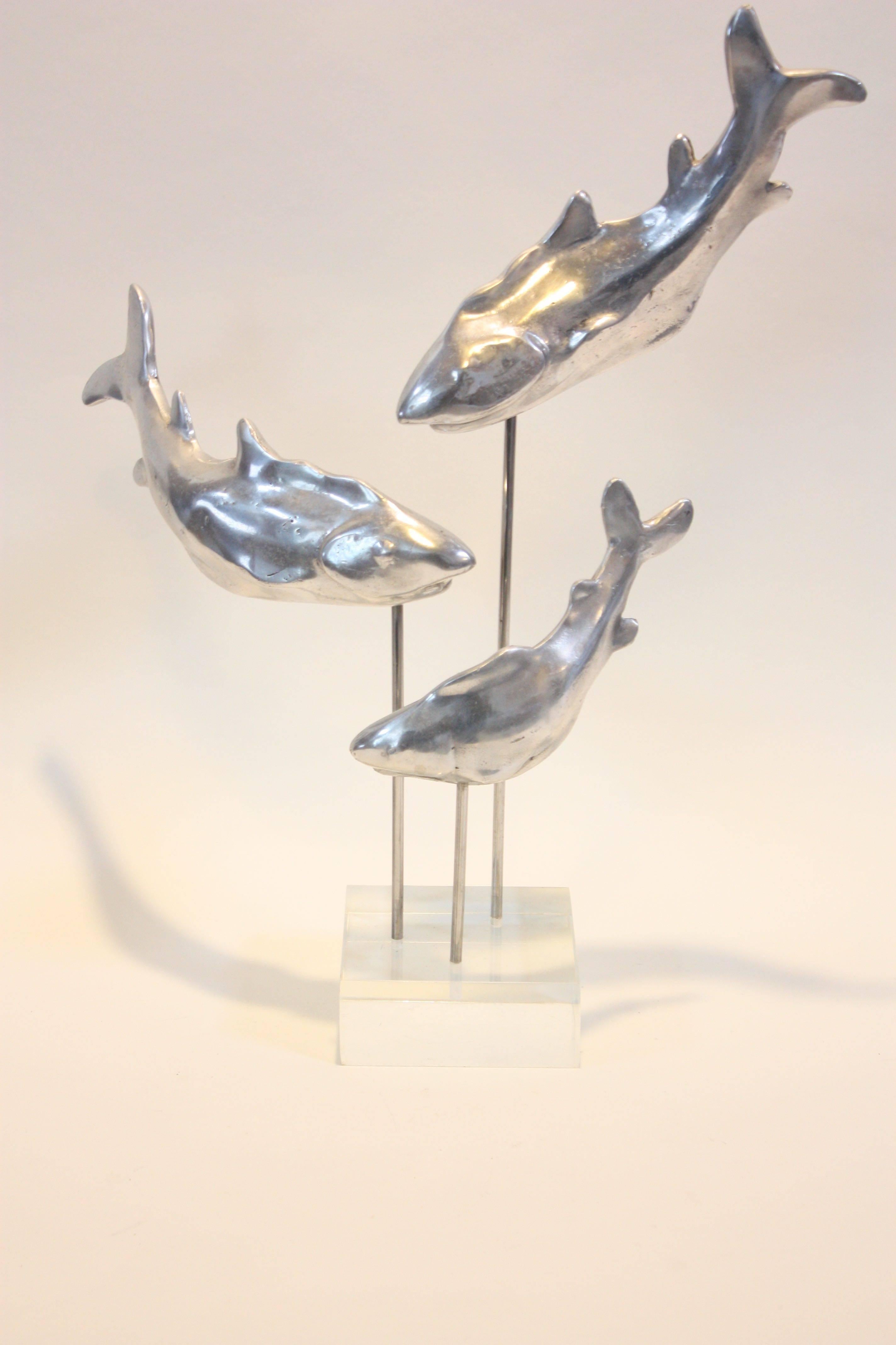 This unique sculpture is composed of three cast-aluminum sharks (representative of a family of sharks given the size disparity between the three) displayed in an ascending spiral formation on rods mounted onto a lucite base. The sharks can removed