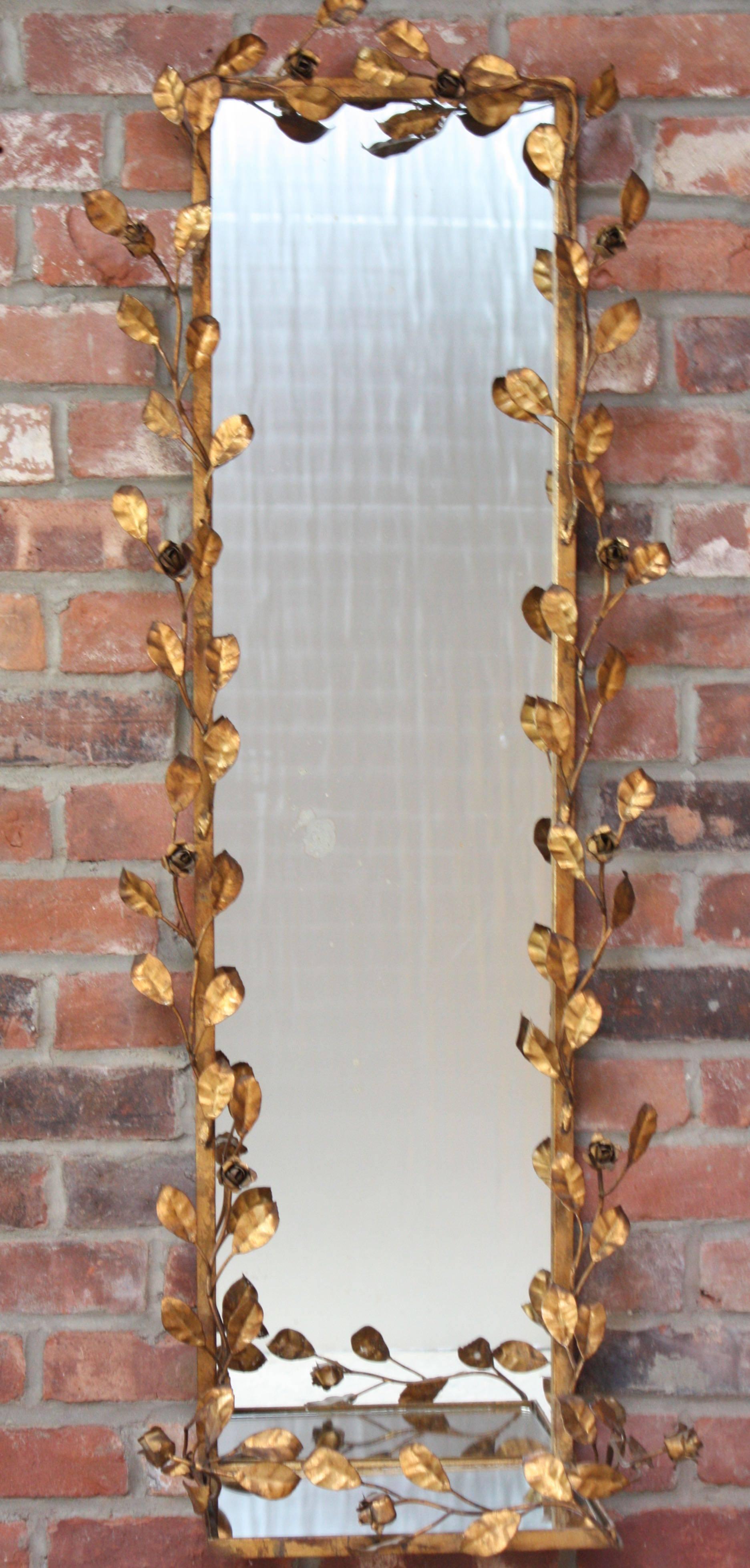 This decorative Italian mirror is adorned by gilt tole flowers and petals. There is rich patina to the gilt from age. There are two pieces of glass in total (the shelf has its own glass panel). There is wear to the glass, as pictured, from dispersed