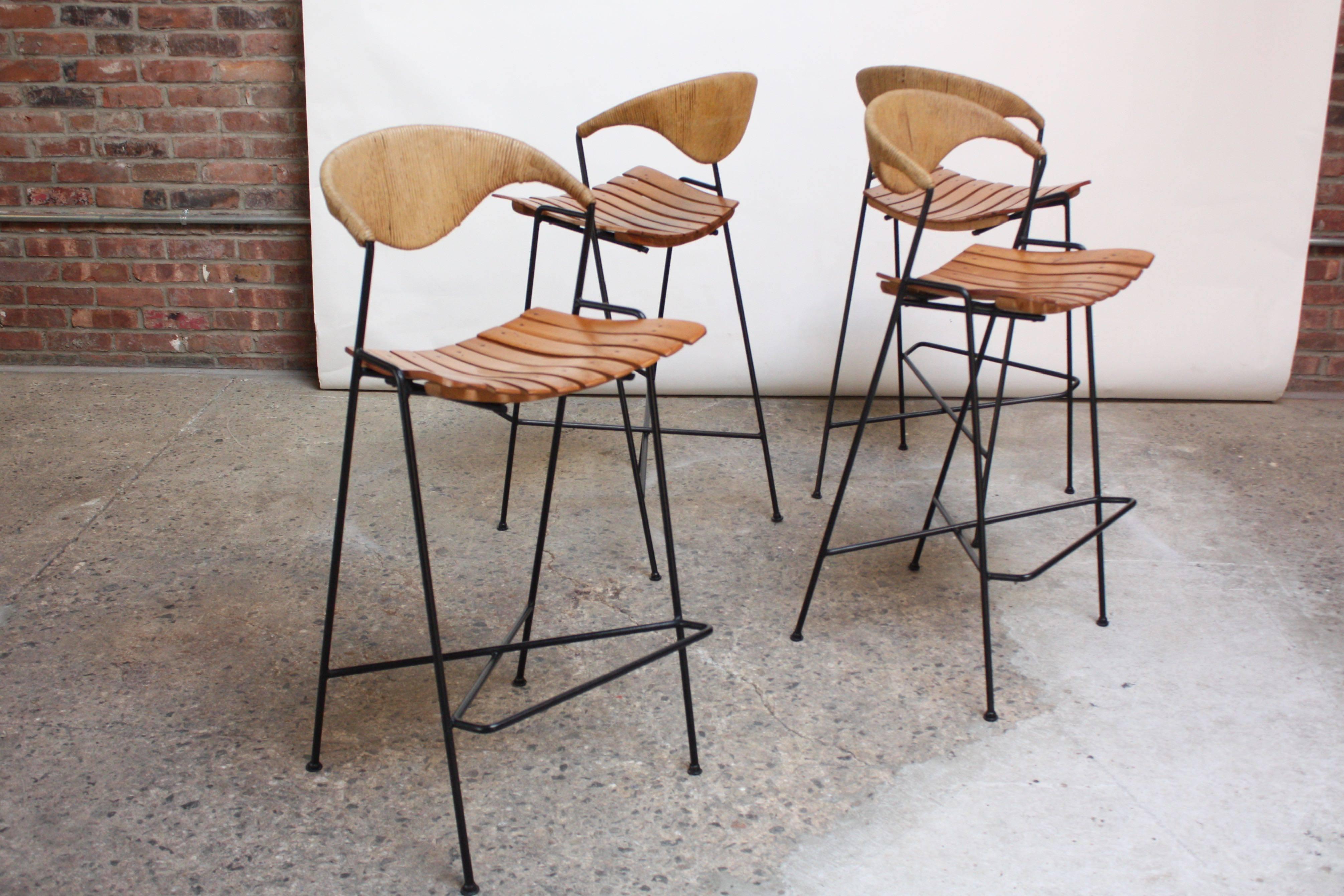 These Umanoff stools are composed of wrought iron frames, stained birch-slatted seats, and rush backs. Sculptural back and unique-form footrest add striking contrast to an otherwise modest form. 
The seat height is 29.5