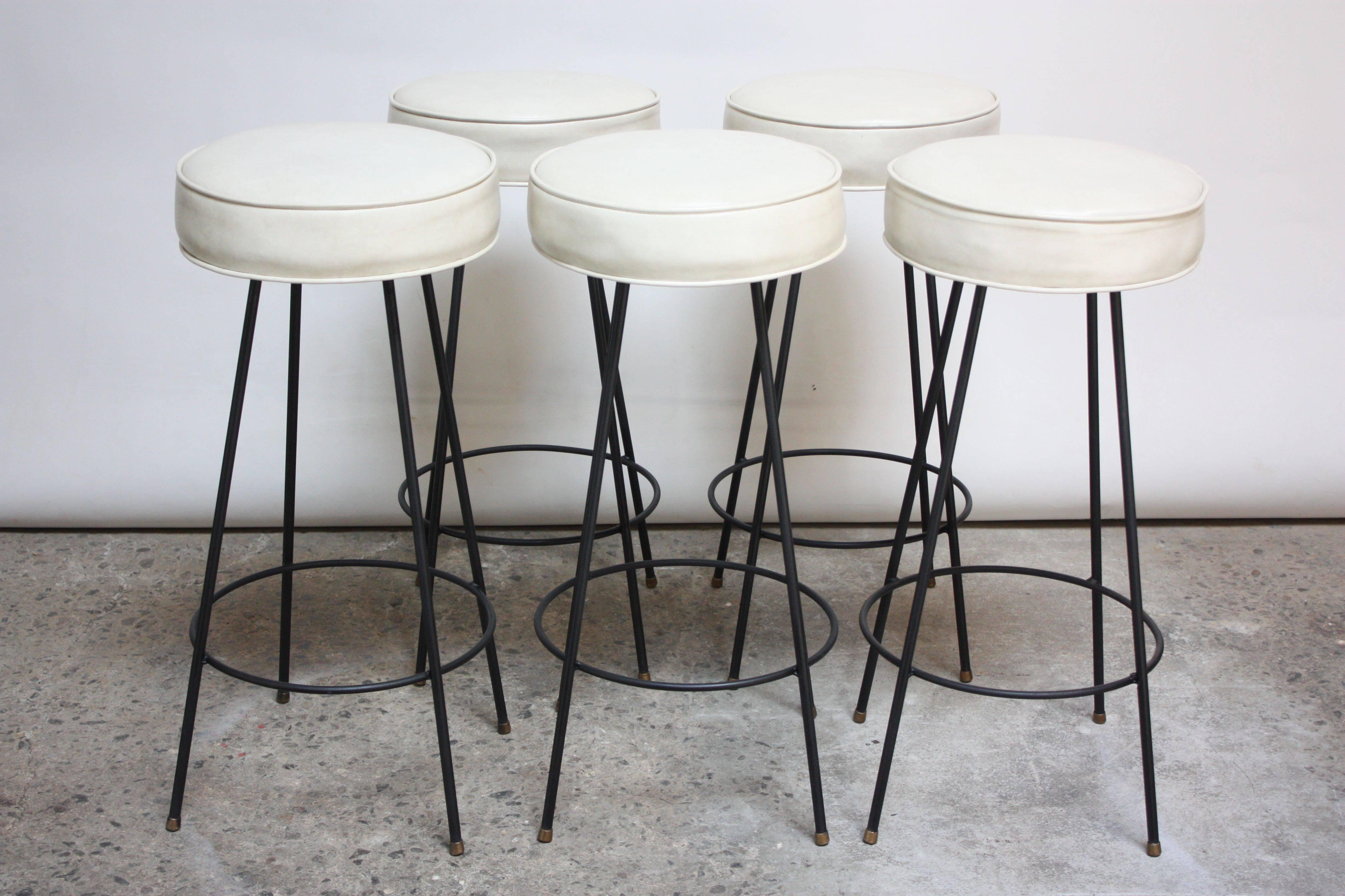 These American modern bar stools are composed of circular vinyl seats supported by four black painted wrought iron legs joined by a circular footrest. The swivel mechanism under the seat allows for full 360 degree rotation. The off-white vinyl is in