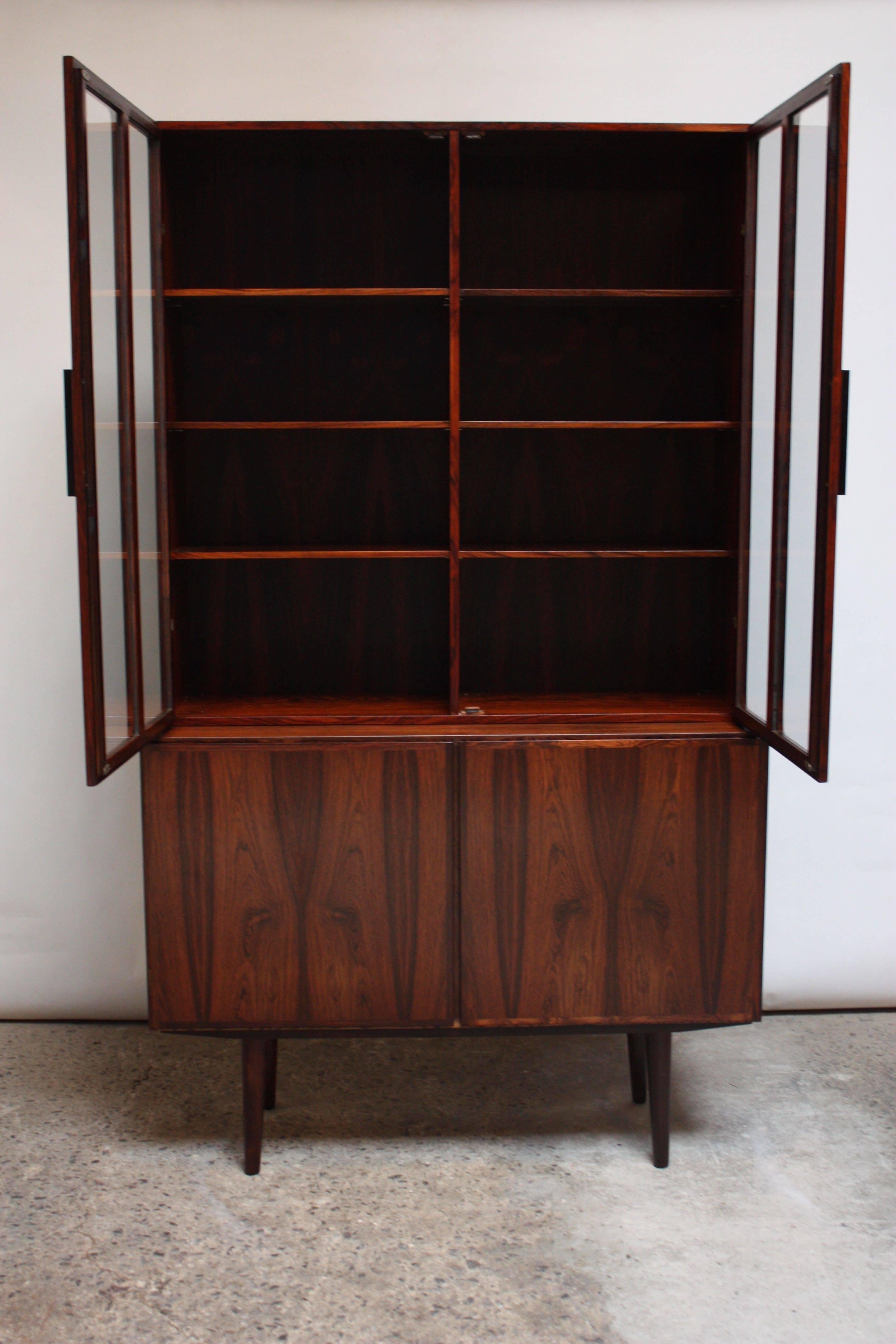 Stunning vitrine with adjustable shelves anchored by a small 2-door cabinet designed by Gunni Omann for Omann Jun. Exquisite bookmatching and vivid rosewood grain. The top case's doors with black metal pulls open to reveal three adjustable shelves /