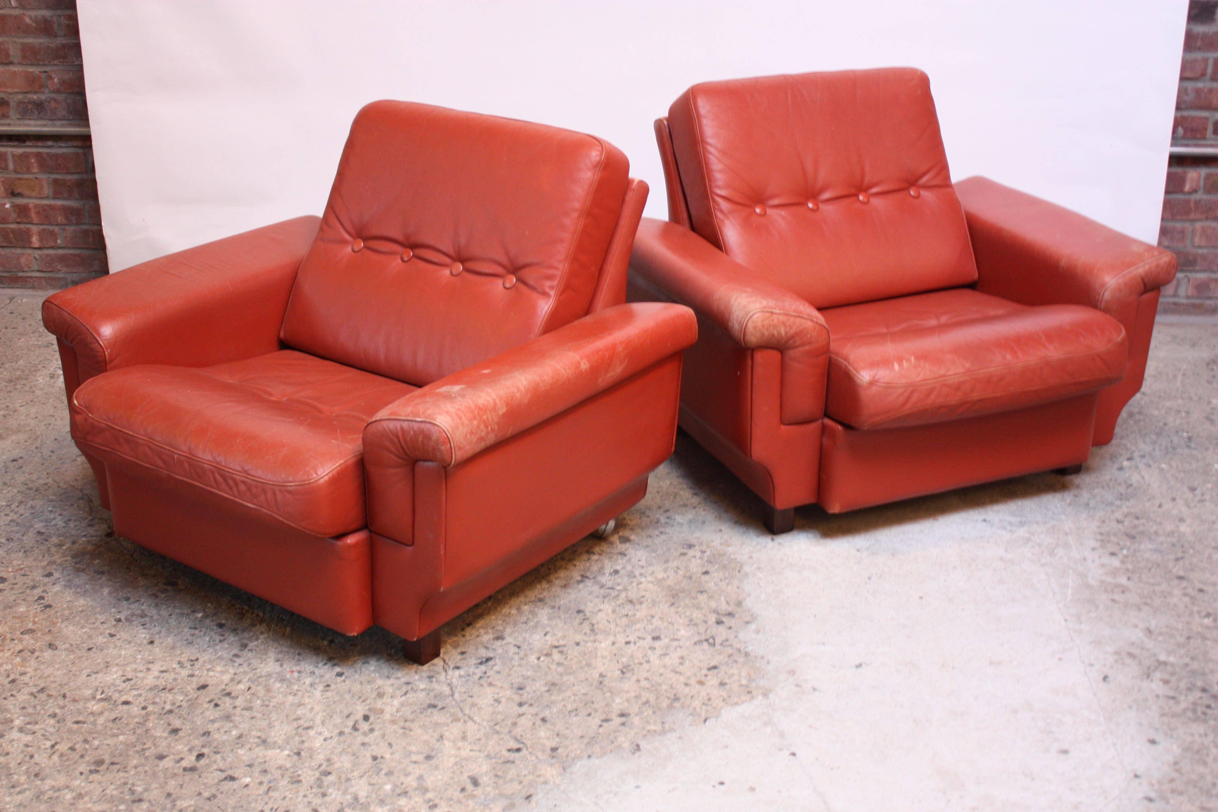 Nicely worn leather lounge chairs in dark coral (Denmark, circa 1970s) composed of two stained mahogany 'block' feet in the front and two caster wheels in the back ('wheelbarrow' design allows for easy mobility).
Leather is nicely distressed with