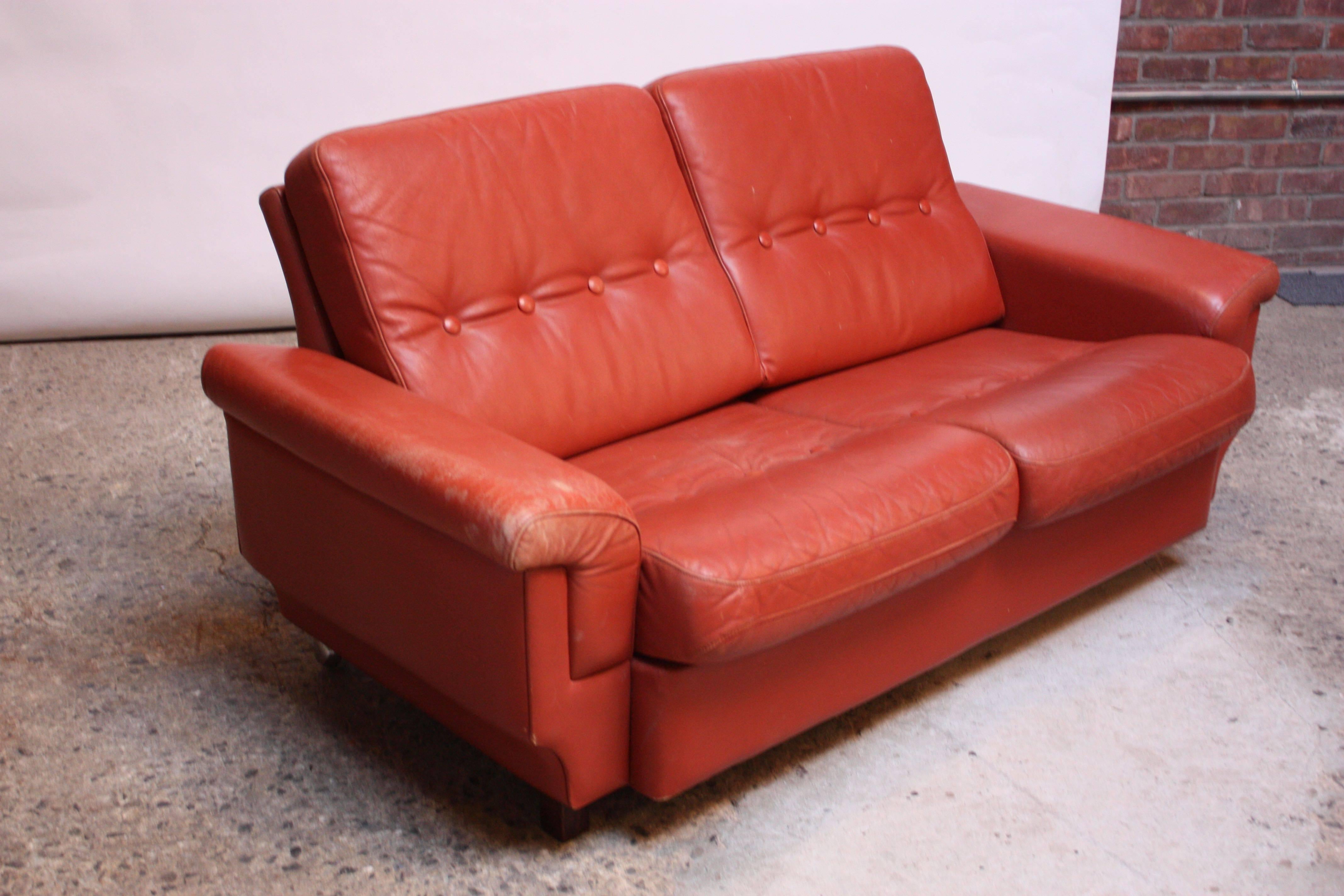 Nicely worn leather loveseat in dark coral (Denmark, circa 1970s) composed of two stained mahogany 'block' feet in the front and two caster wheels in the back ('wheelbarrow' design allows for easy mobility). 
Leather is nicely distressed with spots