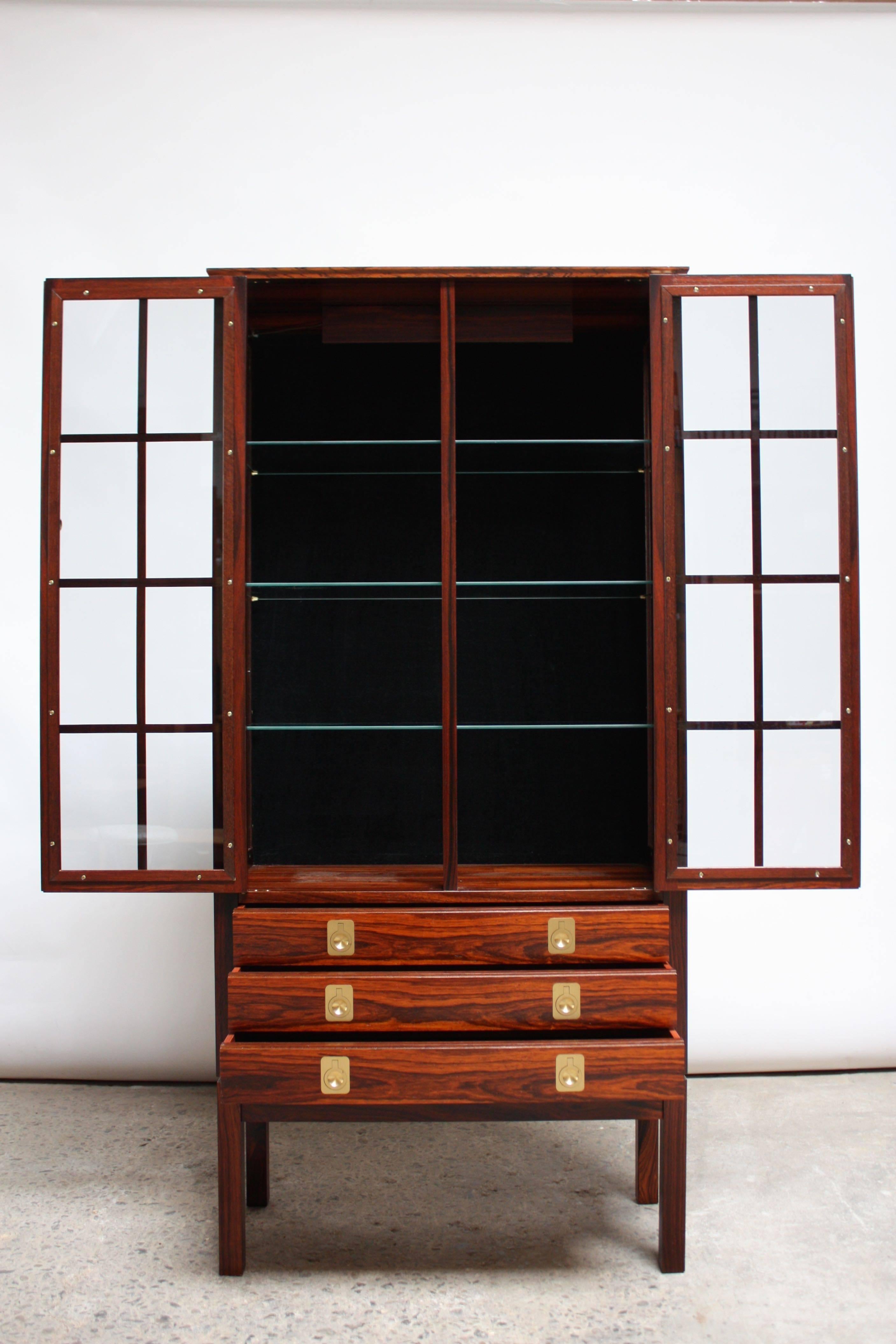 This spectacular curio cabinet was manufactured by Bruksbo of Mellemstrands, Norway in the 1960s and features a working interior light. The glass-fronted doors open to reveal three, non-adjustable glass shelves backed by black, felt lining. The