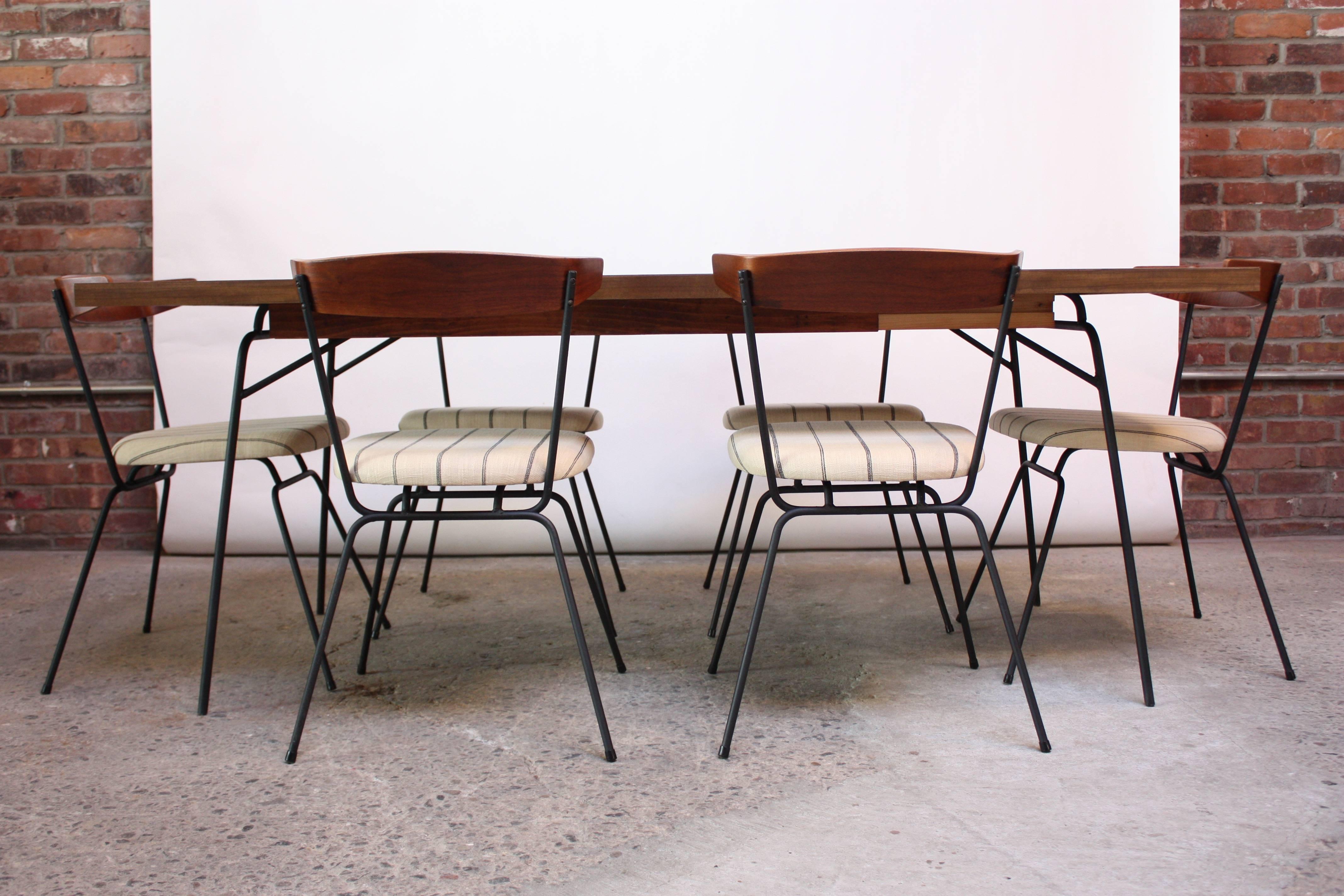 This minimal, elegant dining set was designed in 1951 by Clifford Pascoe for Modernmasters and is comprised of a single-leaf dining table and six chairs, all on hairpin, iron bases. The table features a laminate top 'wrapped' around a solid wood