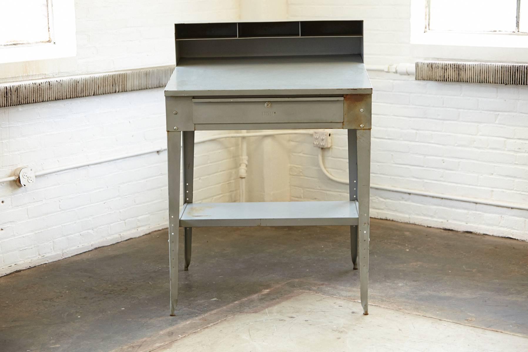 Vintage four-legged 'L' angled steel stand up factory foreman desk with original grey colored powder coat finish. The slanted tabletop has a large writing surface. Single pull out drawer with recessed handle intact. The compartmentalized riser