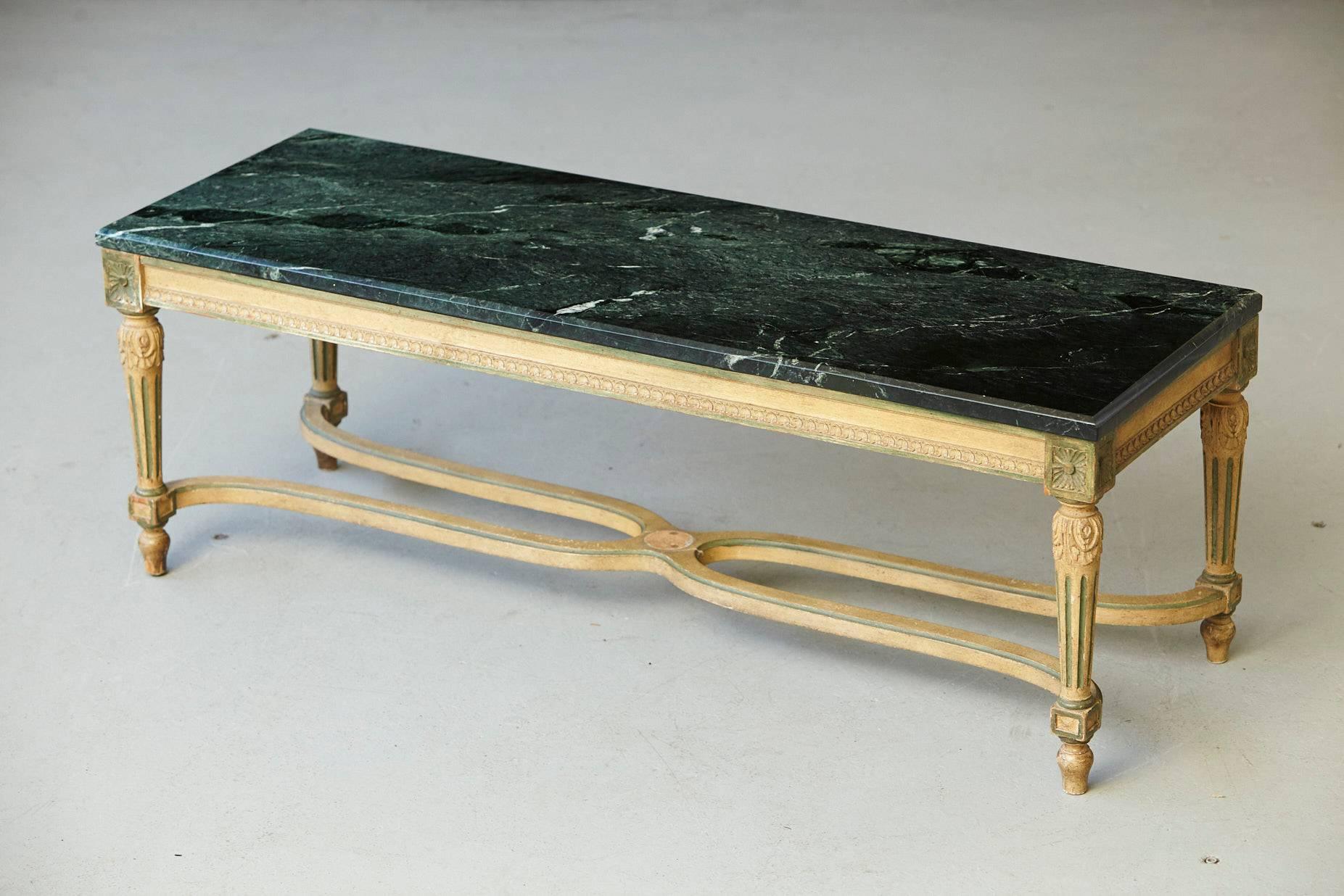 Early 20th century coffee table in a light shimmery gilt paint with marble top in the style of Louis XVI. 
Carved wood frame with four legs joined by an intersecting stretcher, decorative rosette on the stretcher is missing.
The green/black marble