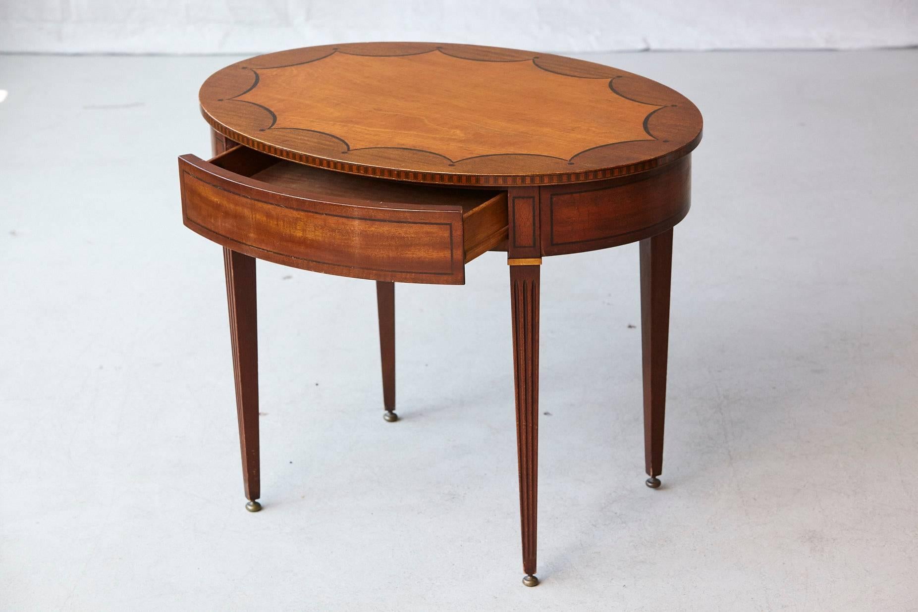 Oval side table with one hidden drawer and graphic marquetry around the apron and the top of the table, raised on tapered legs with brass feed. 
The table has a few scratches and dings to the surface and legs, please refer to the detailed photos.