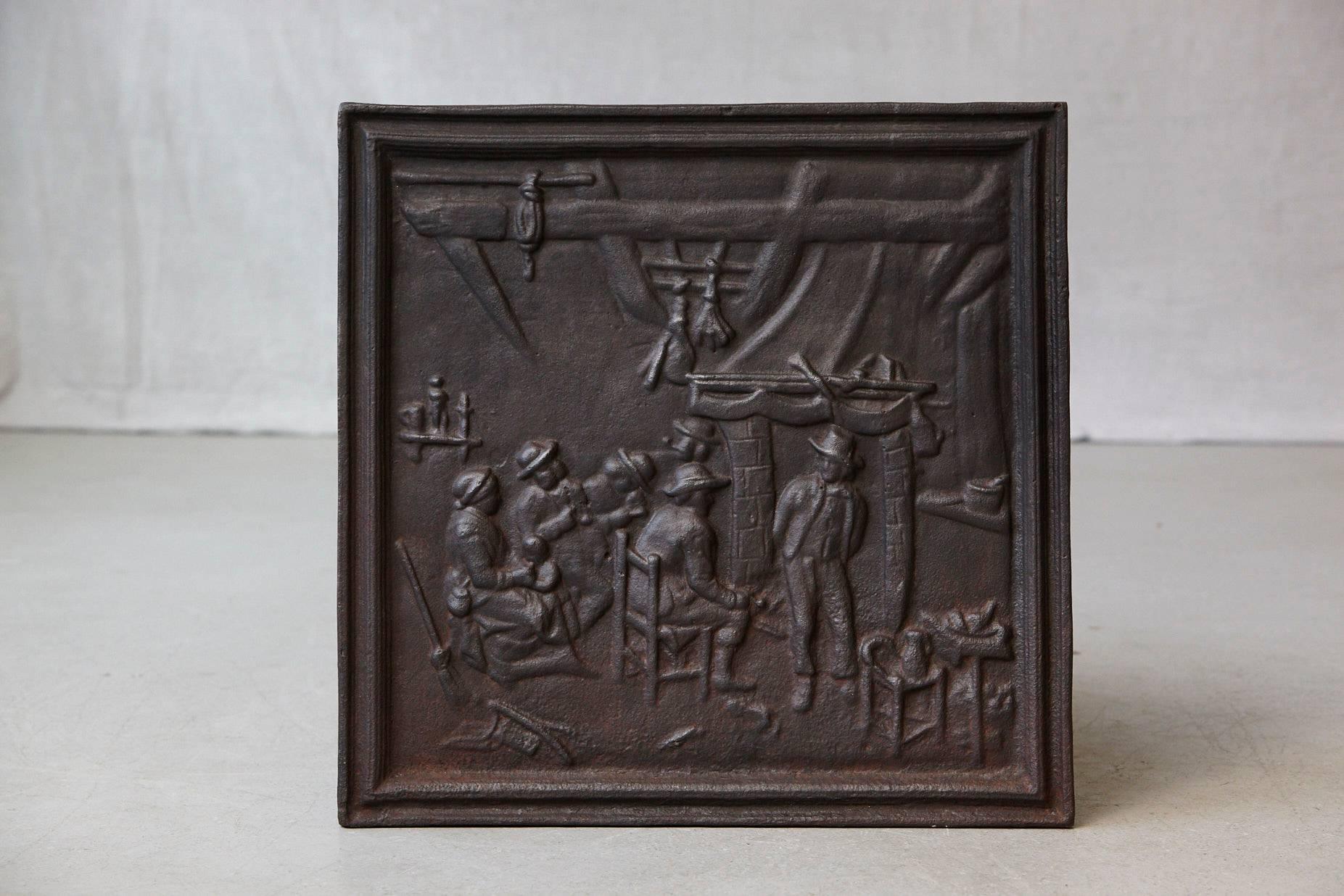 19th century black iron fire back depicting a detailed scene with a group of people in front of a fireplace in a cabin. The large, square fireback is in excellent condition, front and back.