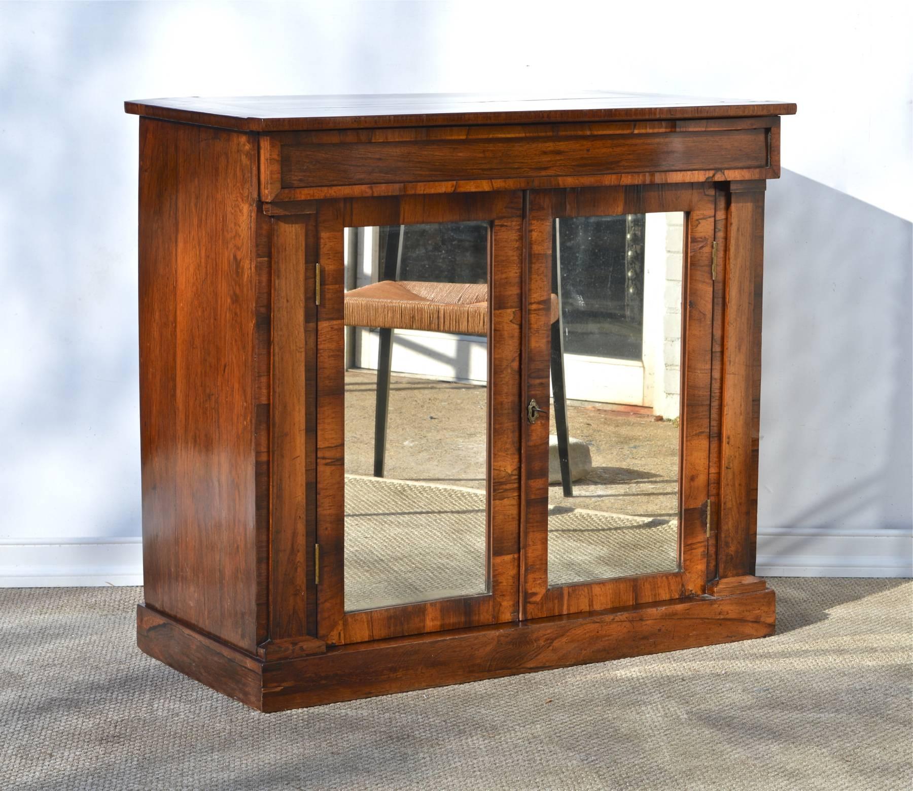 A stunning two door Regency styled bar cabinet of rosewood having a single long sliding drawer for storage above two hinged doors that have been fitted with glamorous mirror glass. The simple case piece shows a gorgeous patina and high polish