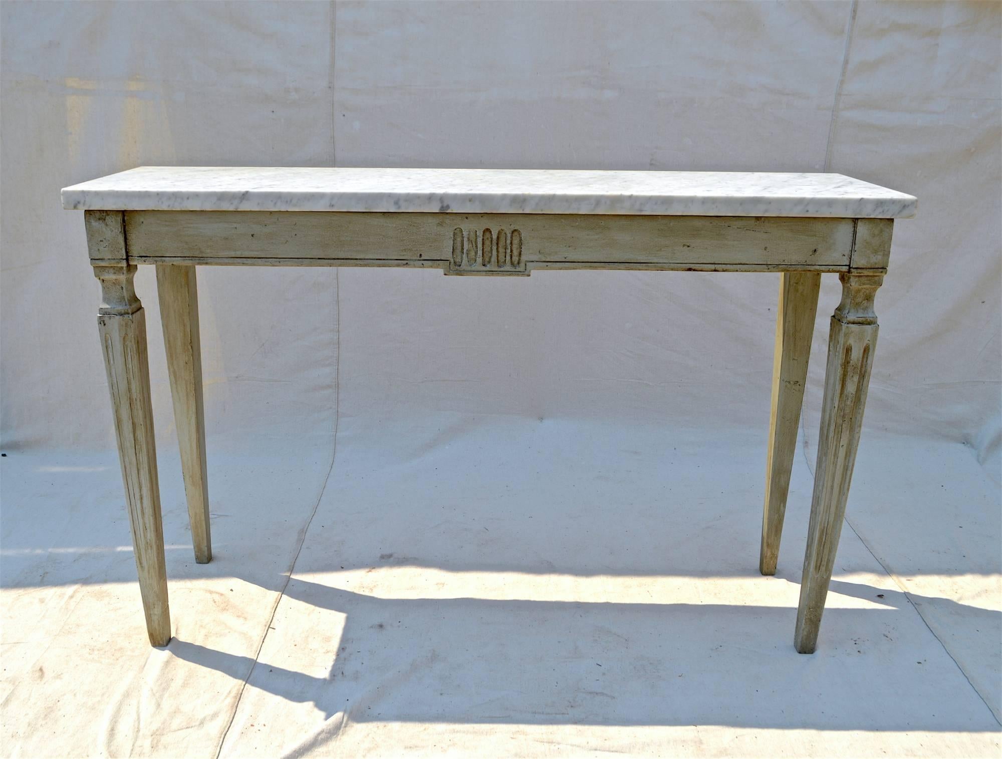 A simple and classic French styled marble top console table in the Neoclassical taste. Warm toned Carrara marble rest upon a rectangular apron having turned and stop reeded front legs and a strong and supportive tapering back leg. The body of the
