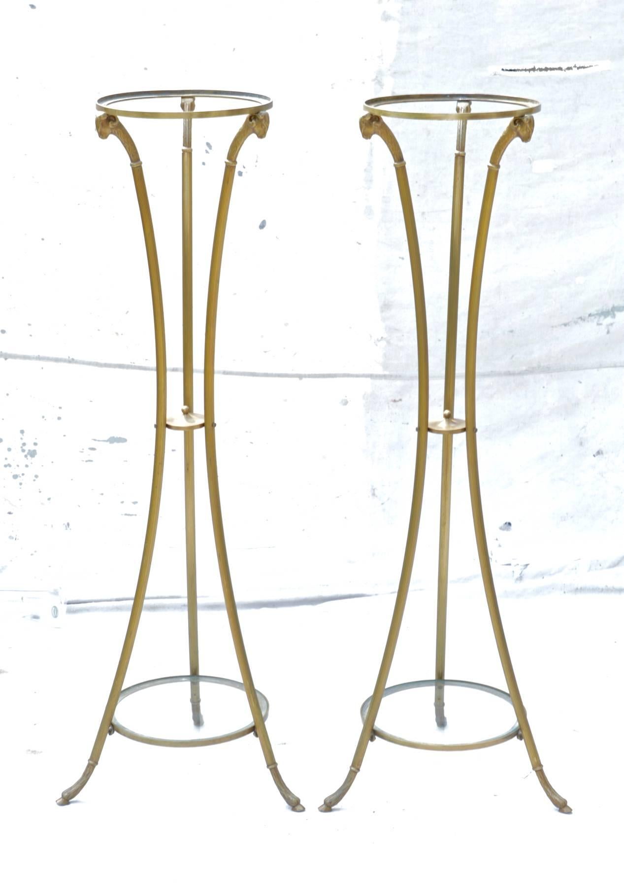 A sleek pair of brass ram's head pedestals in the manner of Maison Jansen, having down swept cylindrical brushed brass legs terminating on hoof feet. Each plant stand has two glass tops from which to decorate. 


 