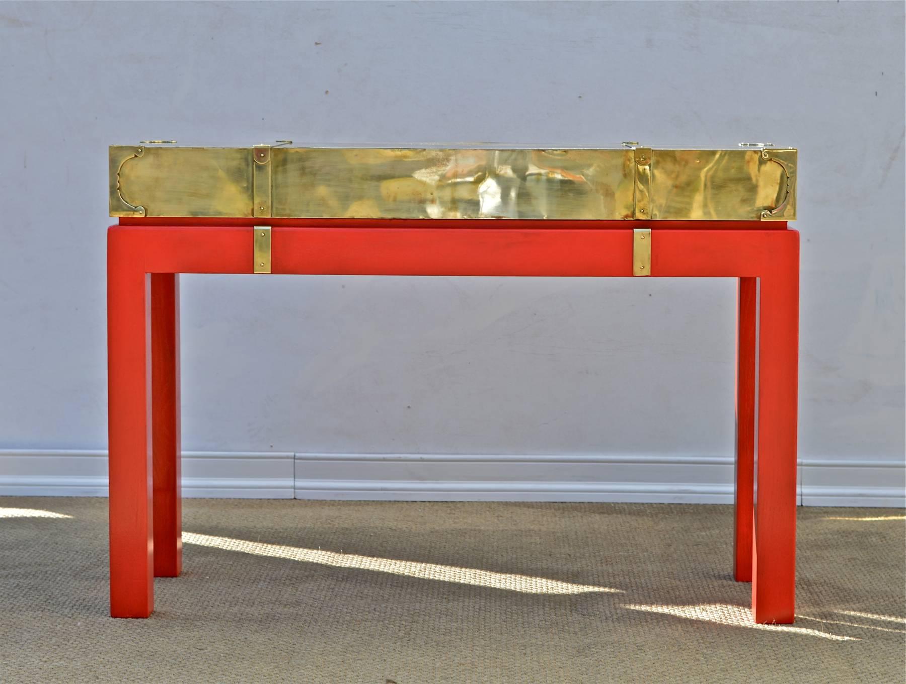 A vintage Campaign style console table in the manner of Karl Springer. The brass wrapped top perches upon a hand enameled oak base of a vibrant red / orange. The matte base juxtapositions well with the glimmer of the brass wrapped top. A decorative