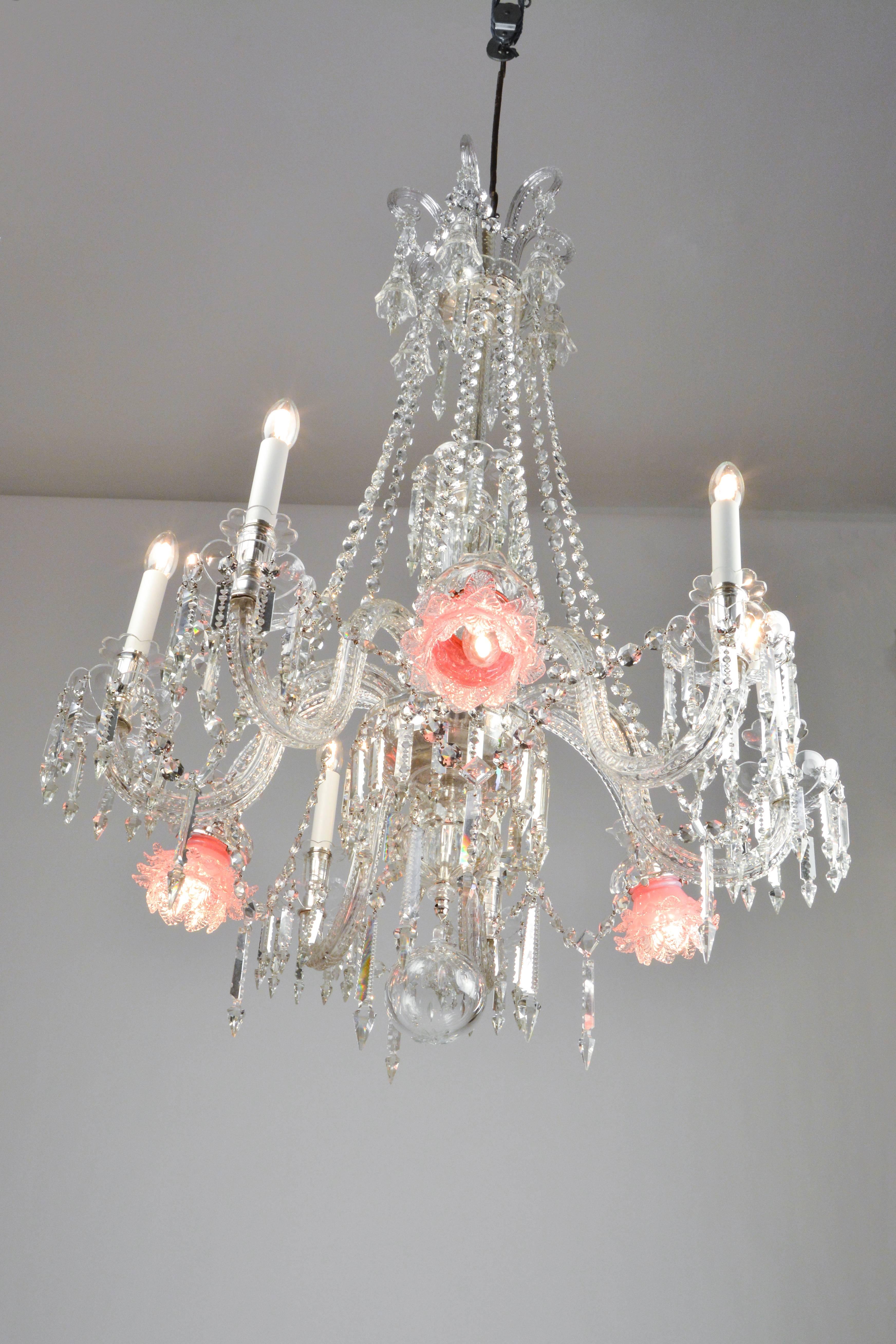 Six-arm chandelier with slender column and tender chains. Extravagant details are the three downward-curved extra arms and their pink flower-shaped glass shades. All arms are extravagantly cut and polished. The crystal sockets on the candles add to