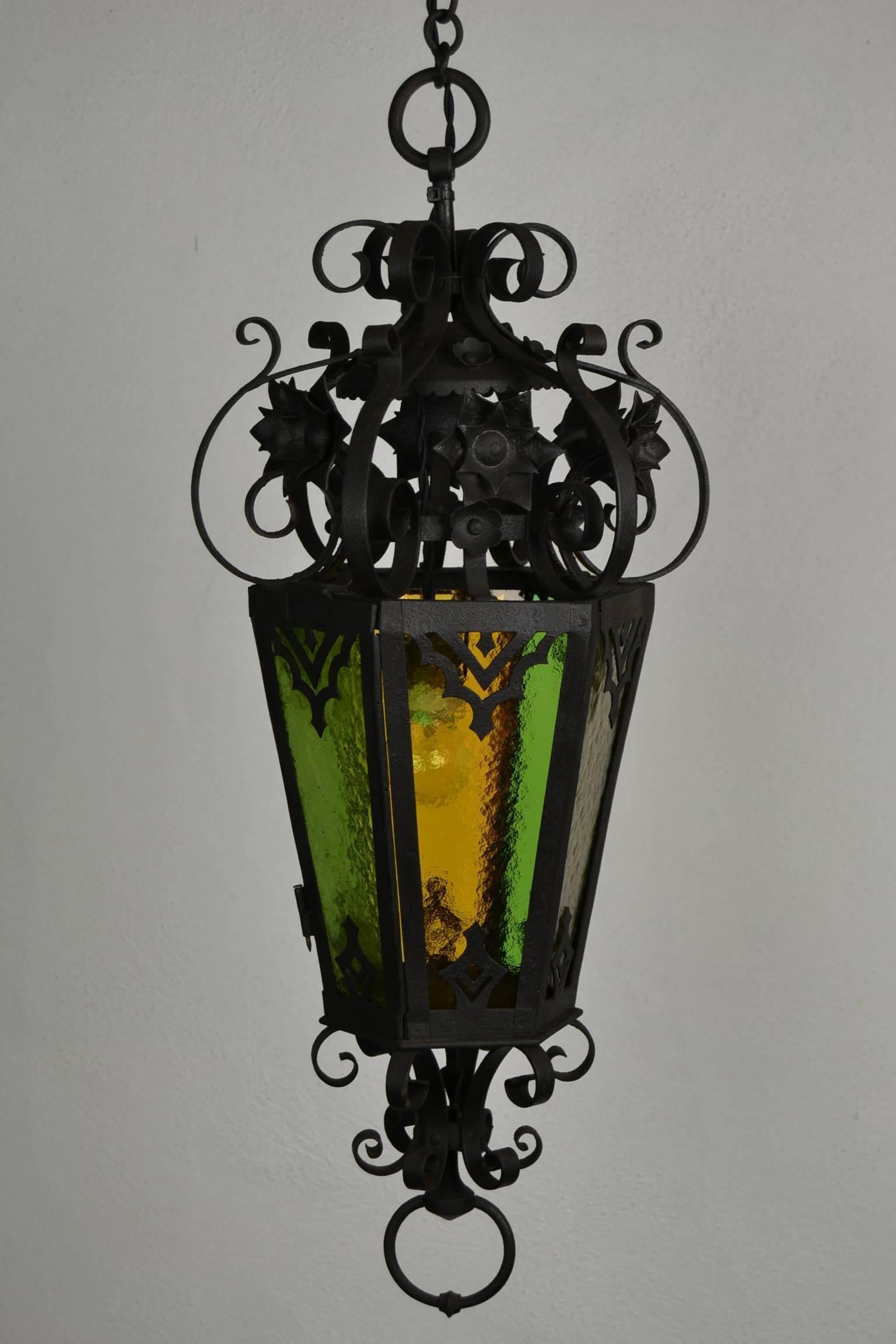 Antique French Wrought Iron Lantern with Colored Glass. 
This Early 20th Century Hexagonal Vestibule Pendant Light is decorated with Flower Ornaments. An Arts and Crafts Lantern Light with 6 Colored Cut glasses: 2 x green, 1 x yellow, 1 rose and 2