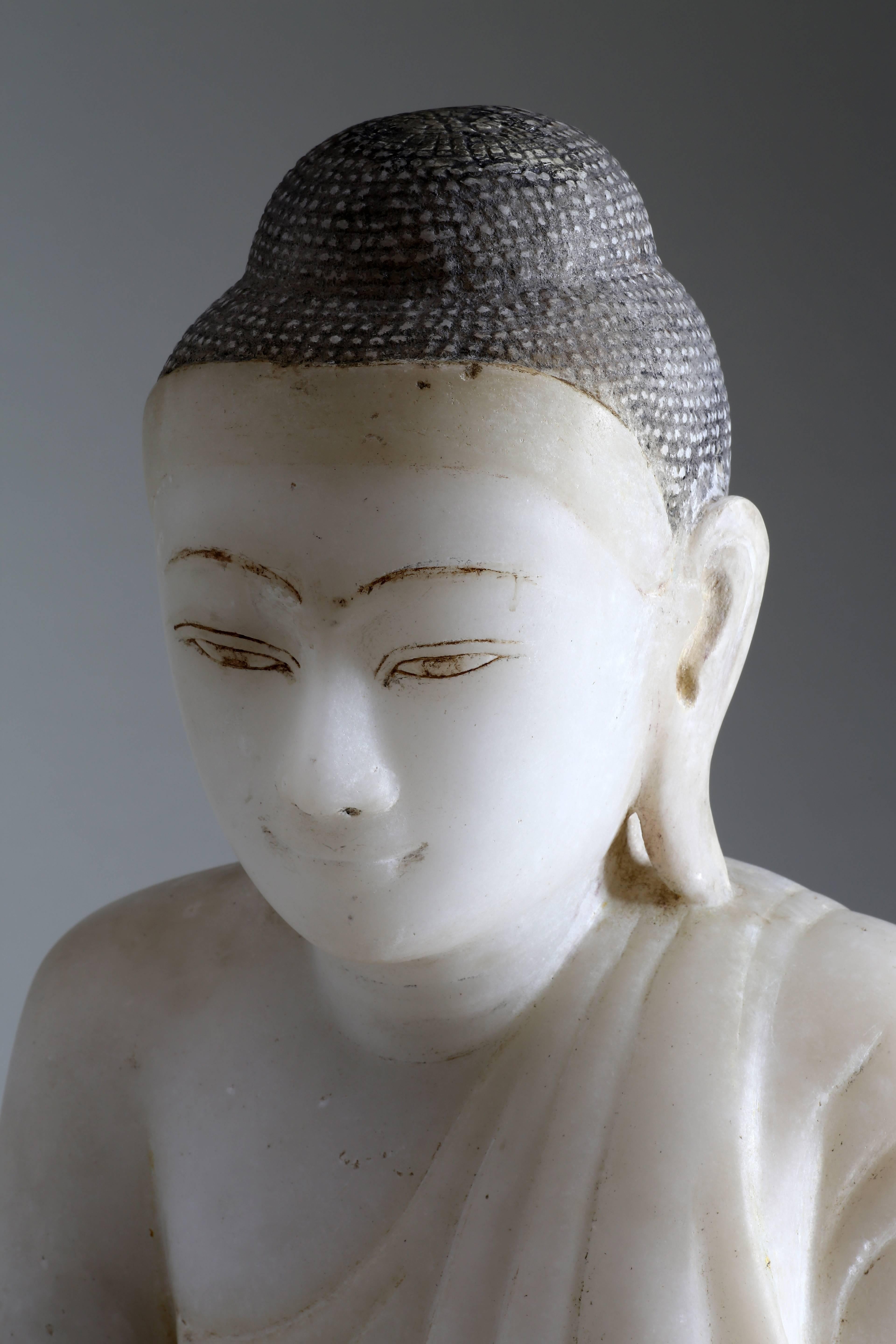 The Buddha is seated on an inscribed low base in vajrasana with bhumisparshamudra, in which the finger-tips of the right hand touch the earth to symbolize the moment of enlightenment. The Buddha is clothed in simple monk’s robes and the billowing