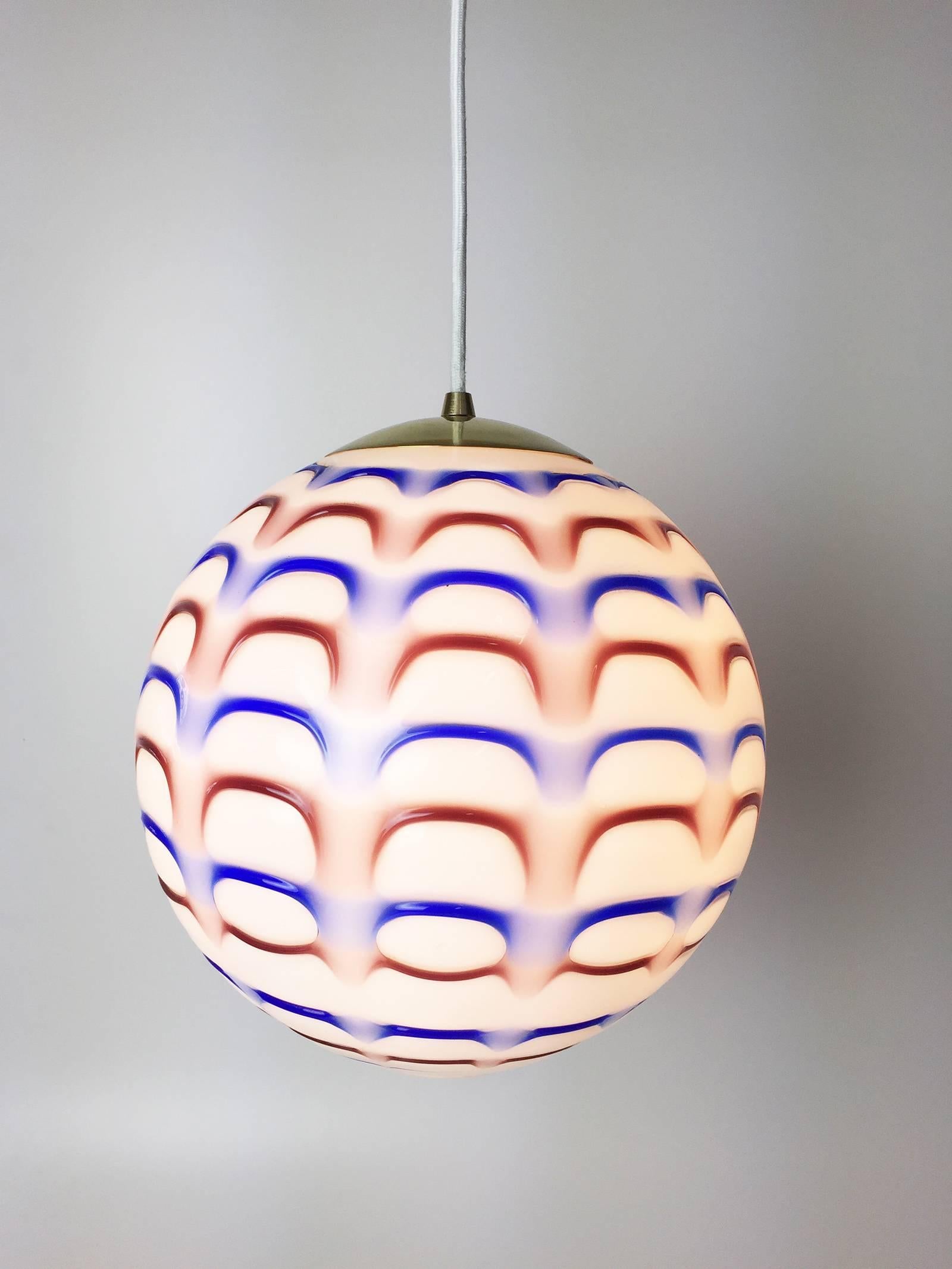 Gorgeous pair of red, white and blue Murano art glass globe pendant lights from the 1960s. Opaque white glass background with red and blue patterning and brass details. Pendants are both currently wired with a 15' white fabric cord with toggle