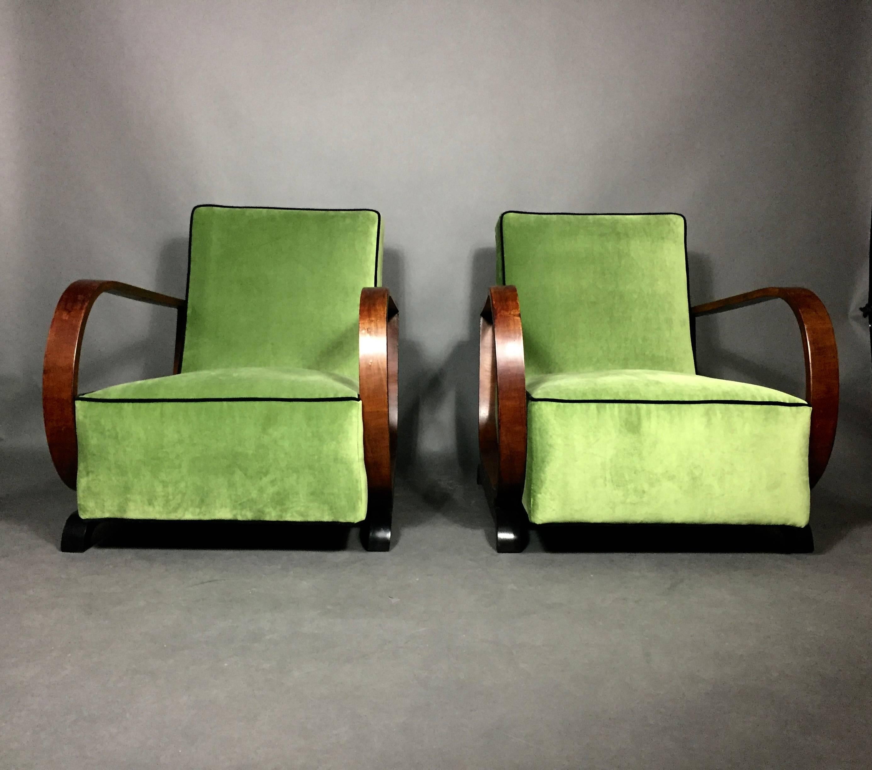 Scale and presence for this pair of low profile Art Deco lounge chairs with Classic curved arms veneered in walnut. New luscious green velvet upholstery with black contrast cording. Bottom black leg/support is a likely renewal. Very minor uneven