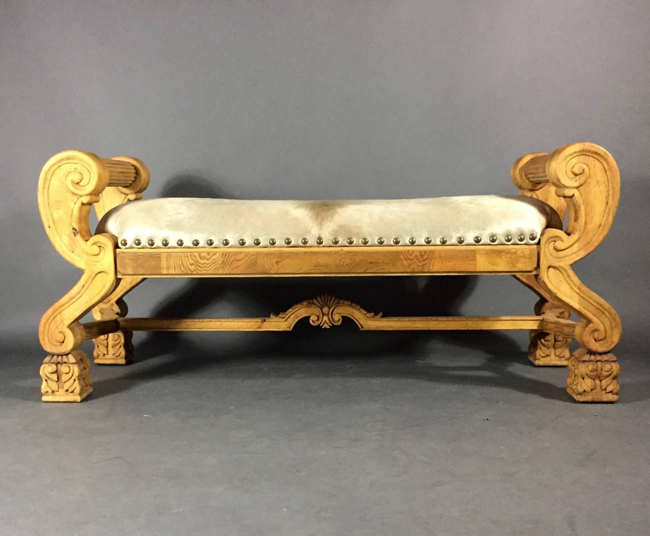 A lively hall or bedroom bench, hand-carved oak with scroll arms in the Empire style and delicately carved base stretcher. New cowhide seating over very solid frame, USA, 20th century. Measures: 53”.