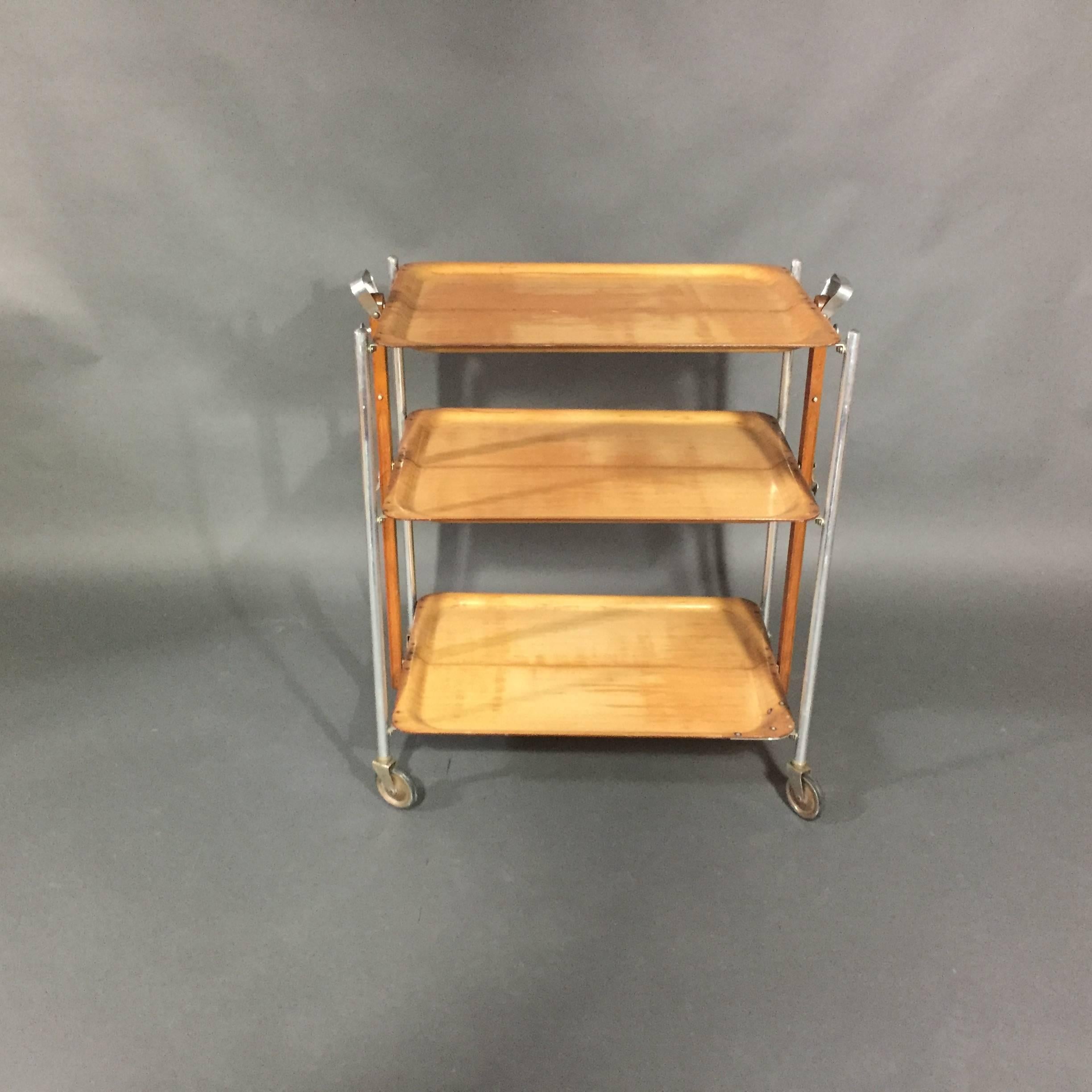 A rather amusing three level bar cart with a structure that allows the entire piece to be folded for easy storage.  Each tray is a molded light wood that is support by chrome legs.  Handy bottle openers to each side.  Labeled "Textable