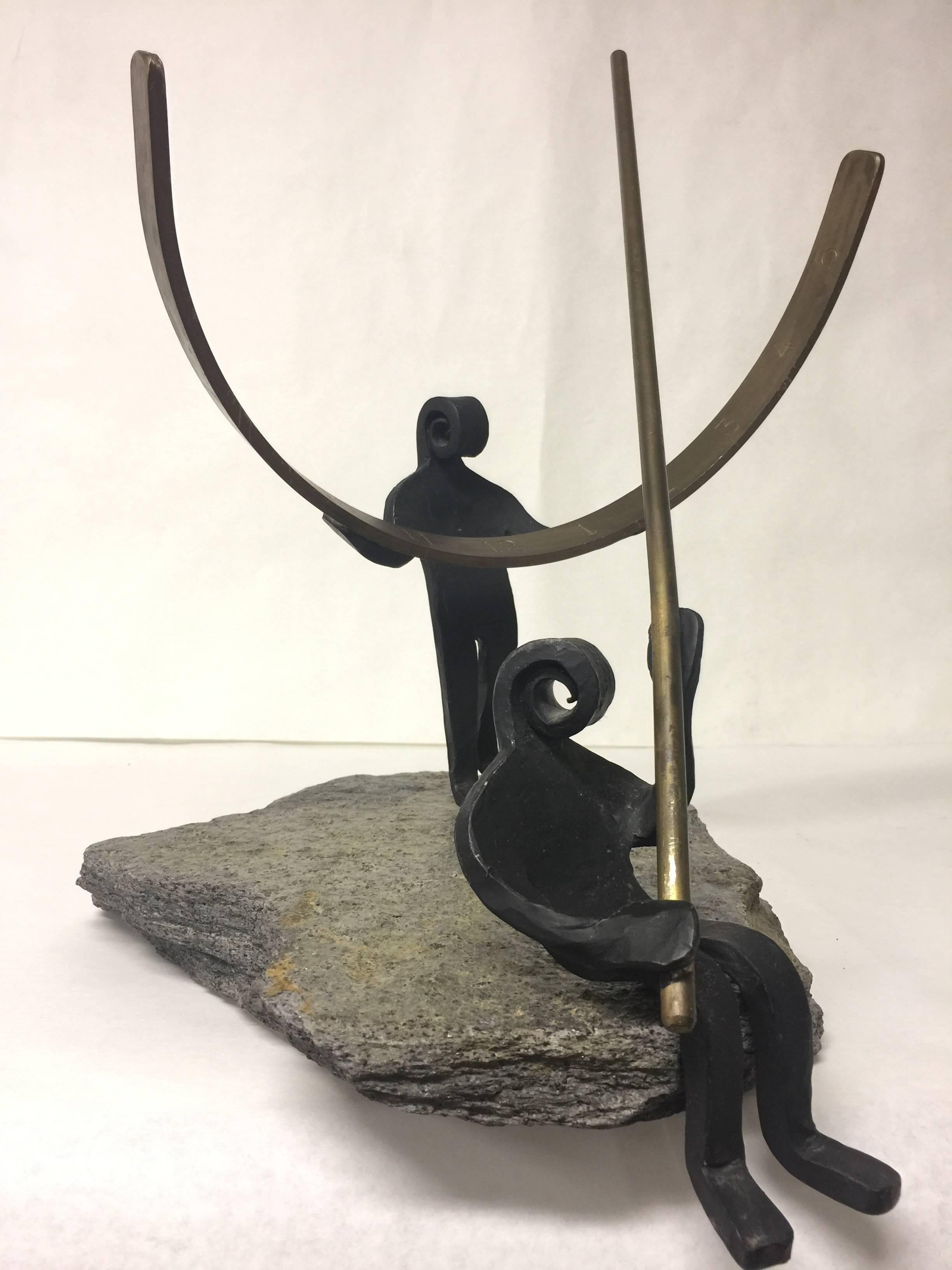 This sundial shows two abstract wrought iron figures, one seated holding the gnomon (the long solid brass rod) and the other standing holding the dial (the curved and incised brass semicircle) both on top of a flat granite like stone. Each figure